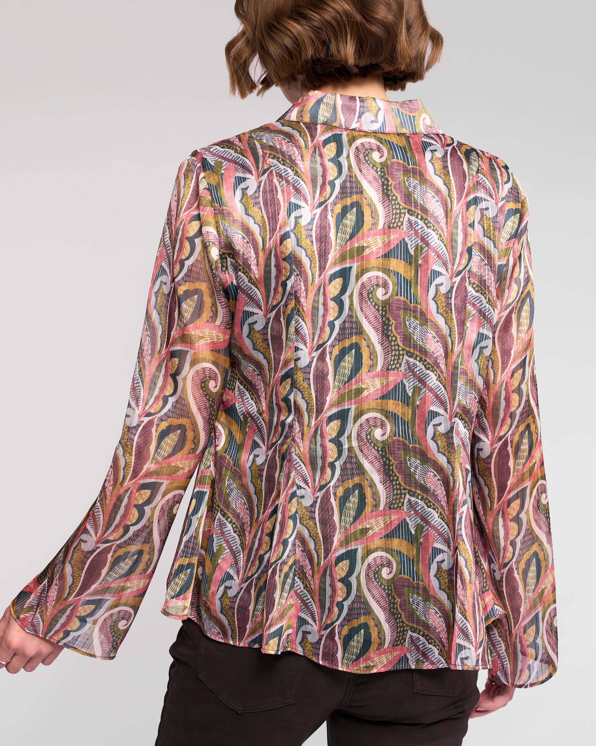 A person with short, wavy brown hair is shown from the back wearing the Vassalli Valley Shirt - Sable, a long-sleeve collared chiffon shirt featuring a colorful paisley-inspired leaf pattern in pink, yellow, blue, and white.