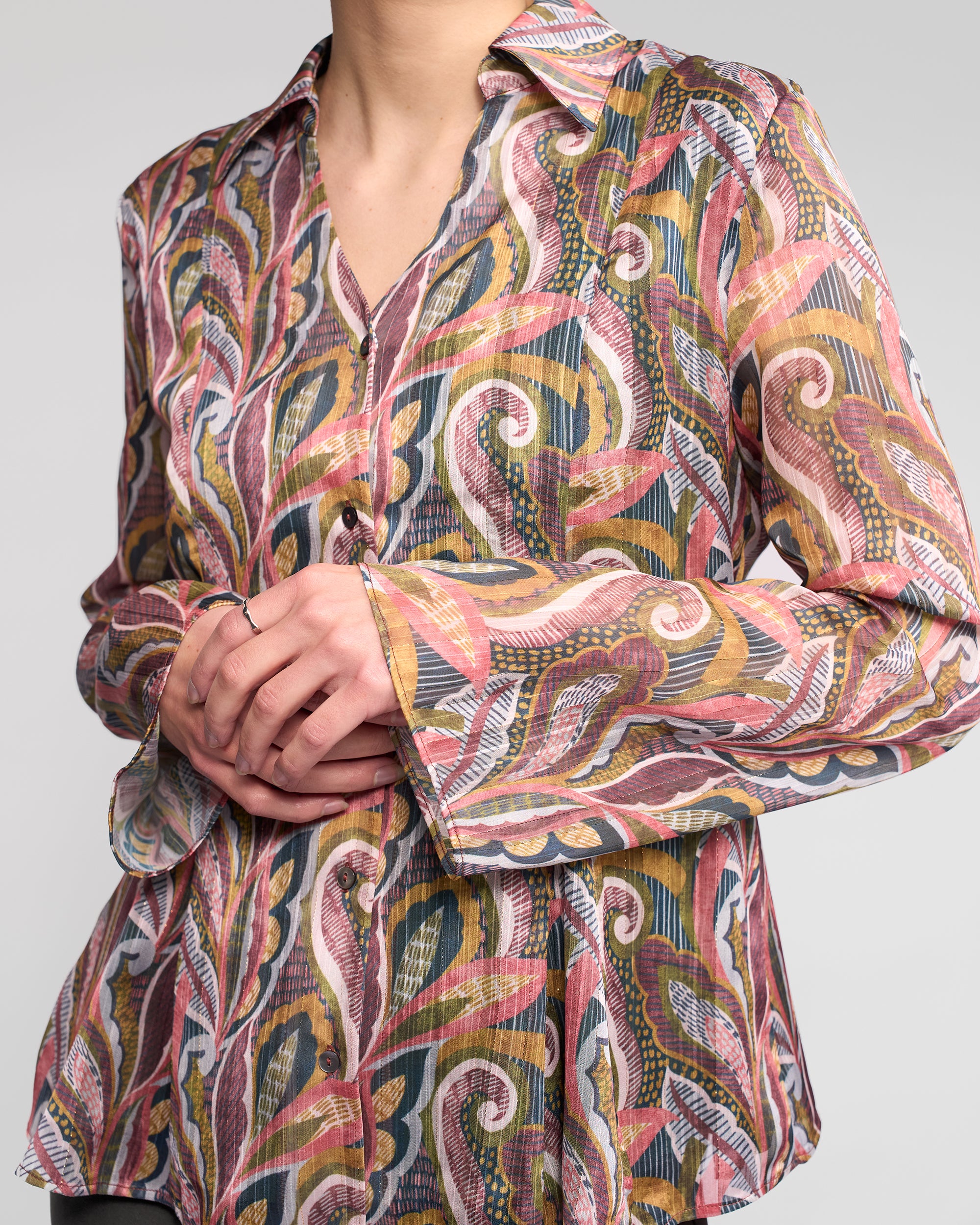 A person wears the Vassalli Valley Shirt - Sable, a long-sleeve chiffon top featuring an abstract, colorful pink, yellow, green, and white design. Their hands are crossed and their face is partially out of frame.