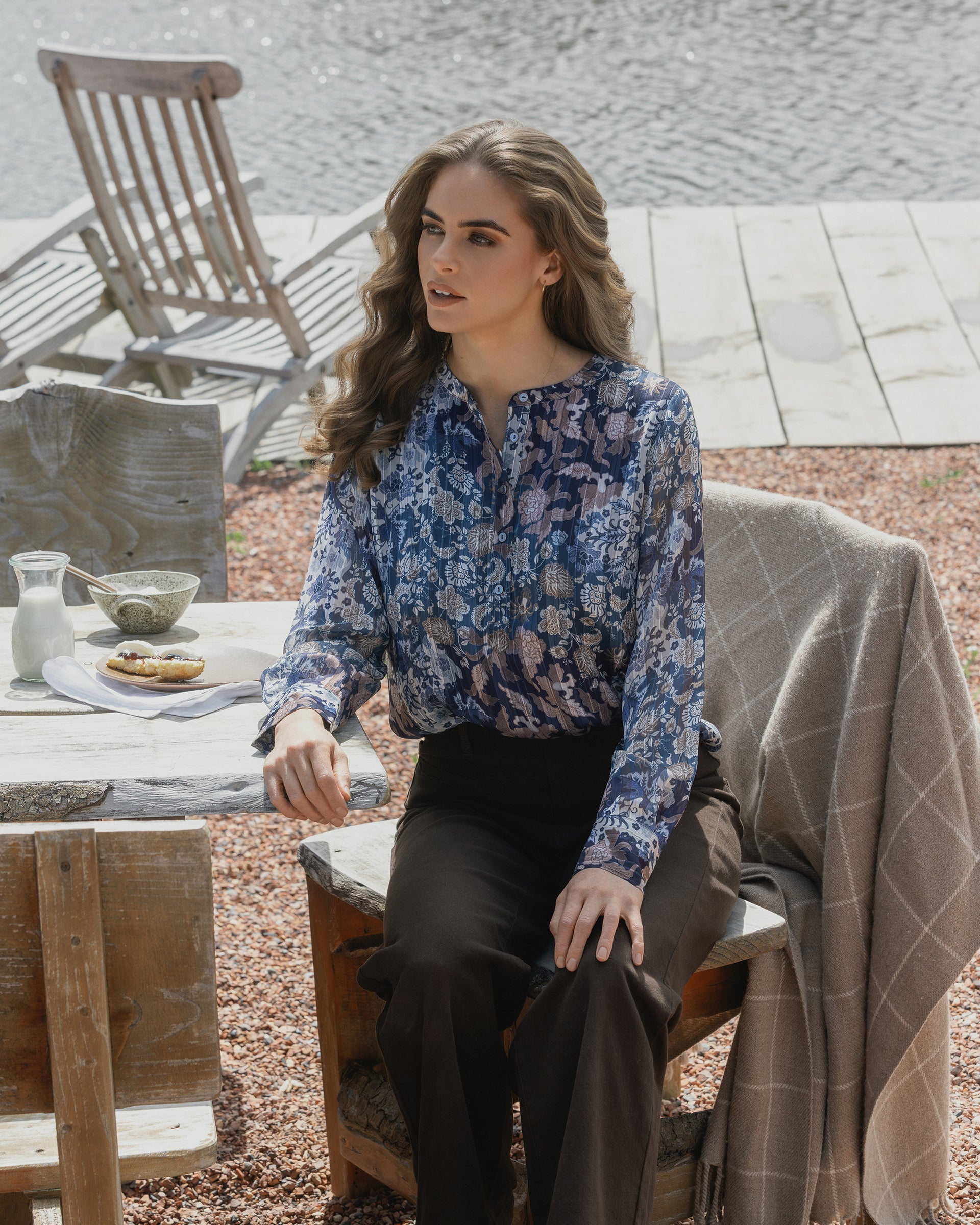 A woman with wavy brown hair, wearing the Vassalli Rhythm Top - Rhea and dark pants, sits at an outdoor lakefront table. Wooden chairs and a dock are behind her; a blanket drapes her chair and food is on the table.