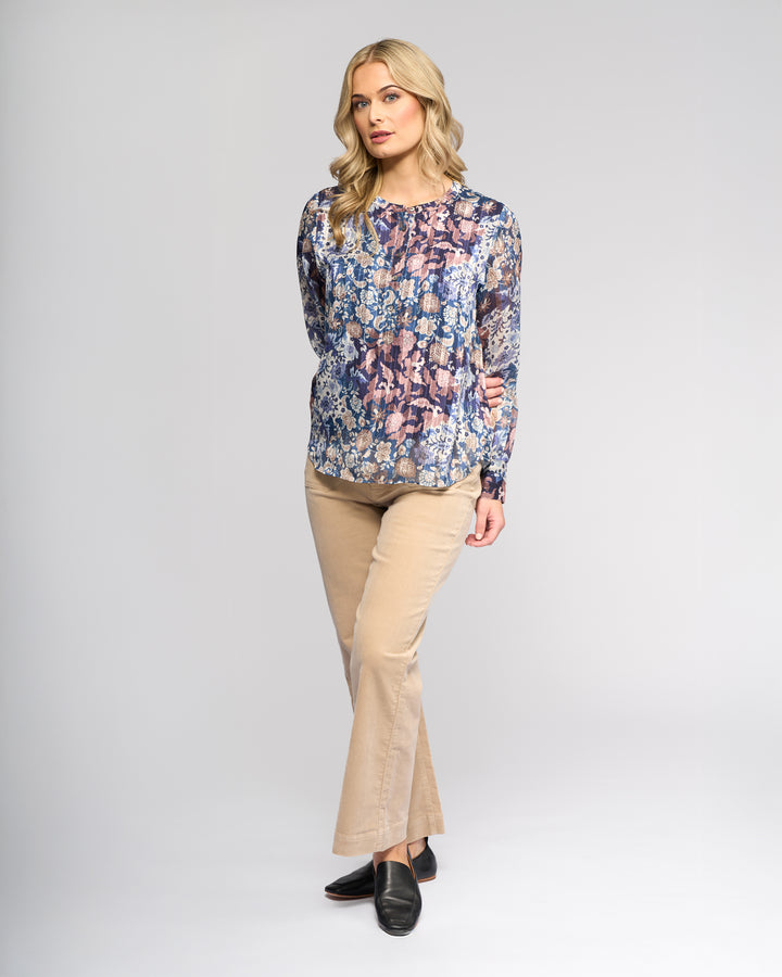 A woman with wavy blonde hair wears the Vassalli Rhythm Top - Rhea, featuring a long-sleeve floral print chiffon design, paired with beige pants and black shoes, standing against a plain light background.