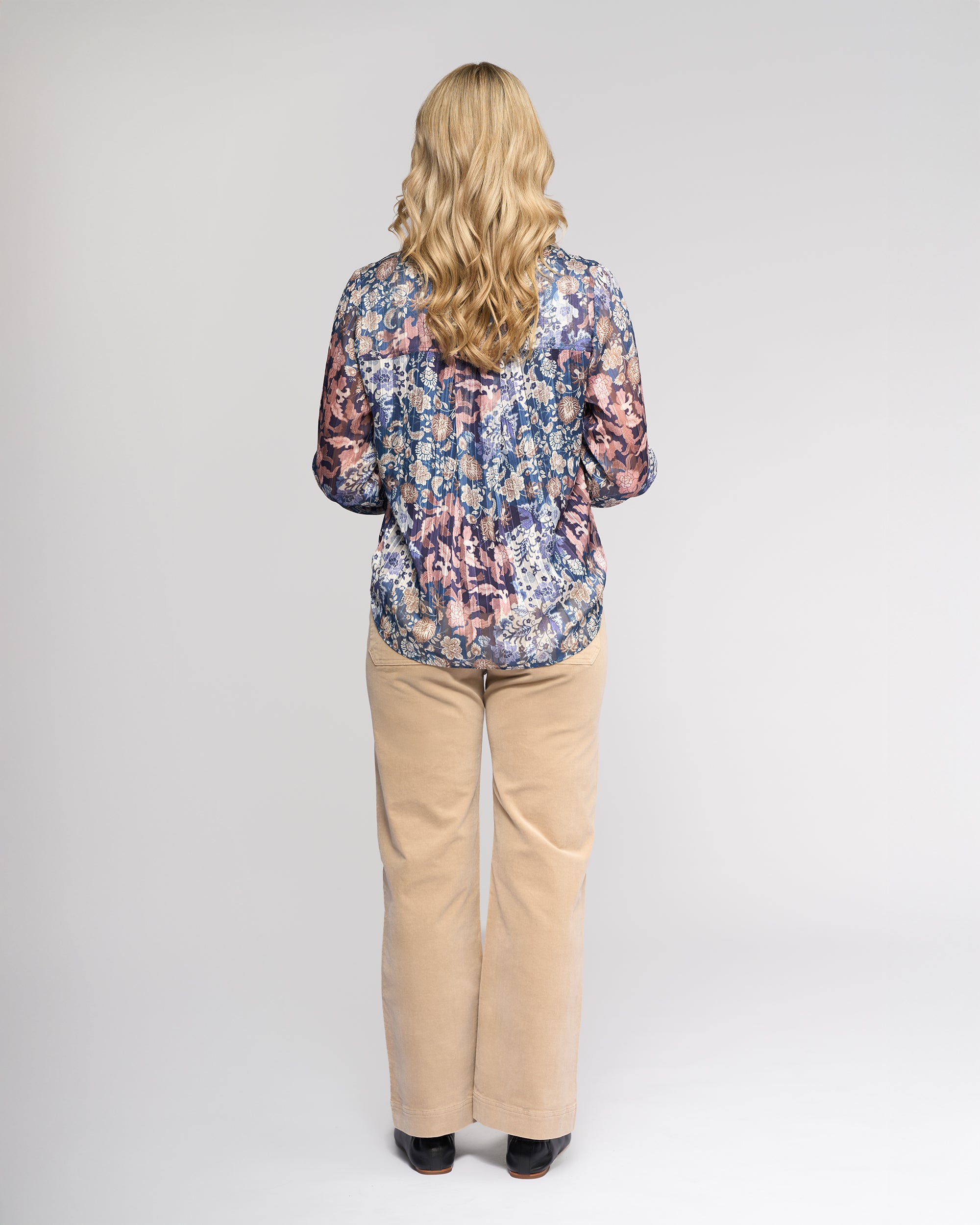 A person with long blonde hair stands facing away, wearing the Vassalli Rhythm Top - Rhea, a floral print long-sleeve chiffon shirt, paired with beige pants against a plain light background.