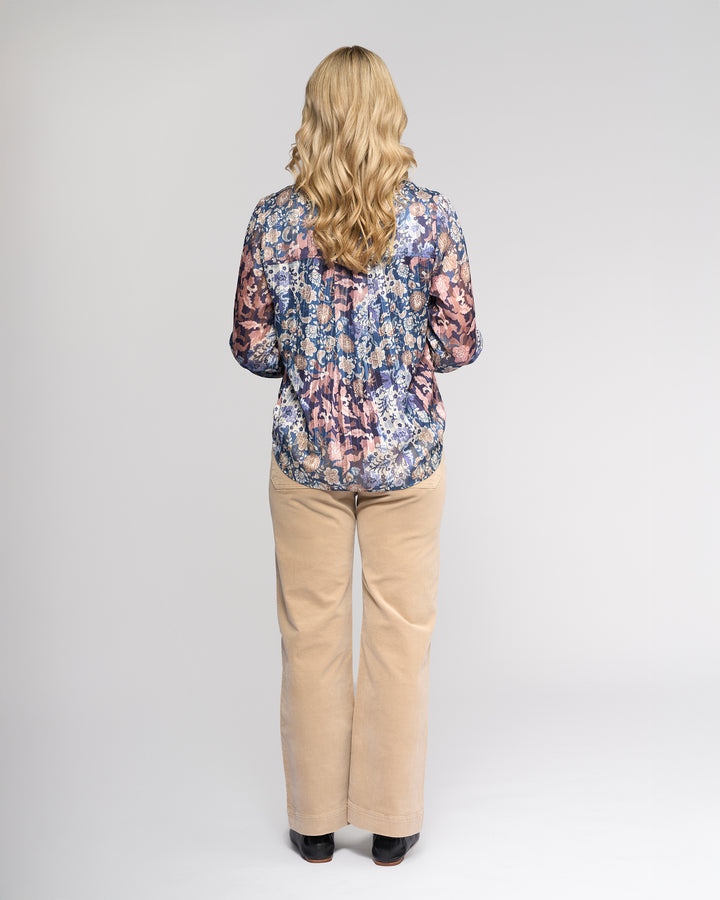 A person with long blonde hair stands facing away, wearing the Vassalli Rhythm Top - Rhea, a floral print long-sleeve chiffon shirt, paired with beige pants against a plain light background.
