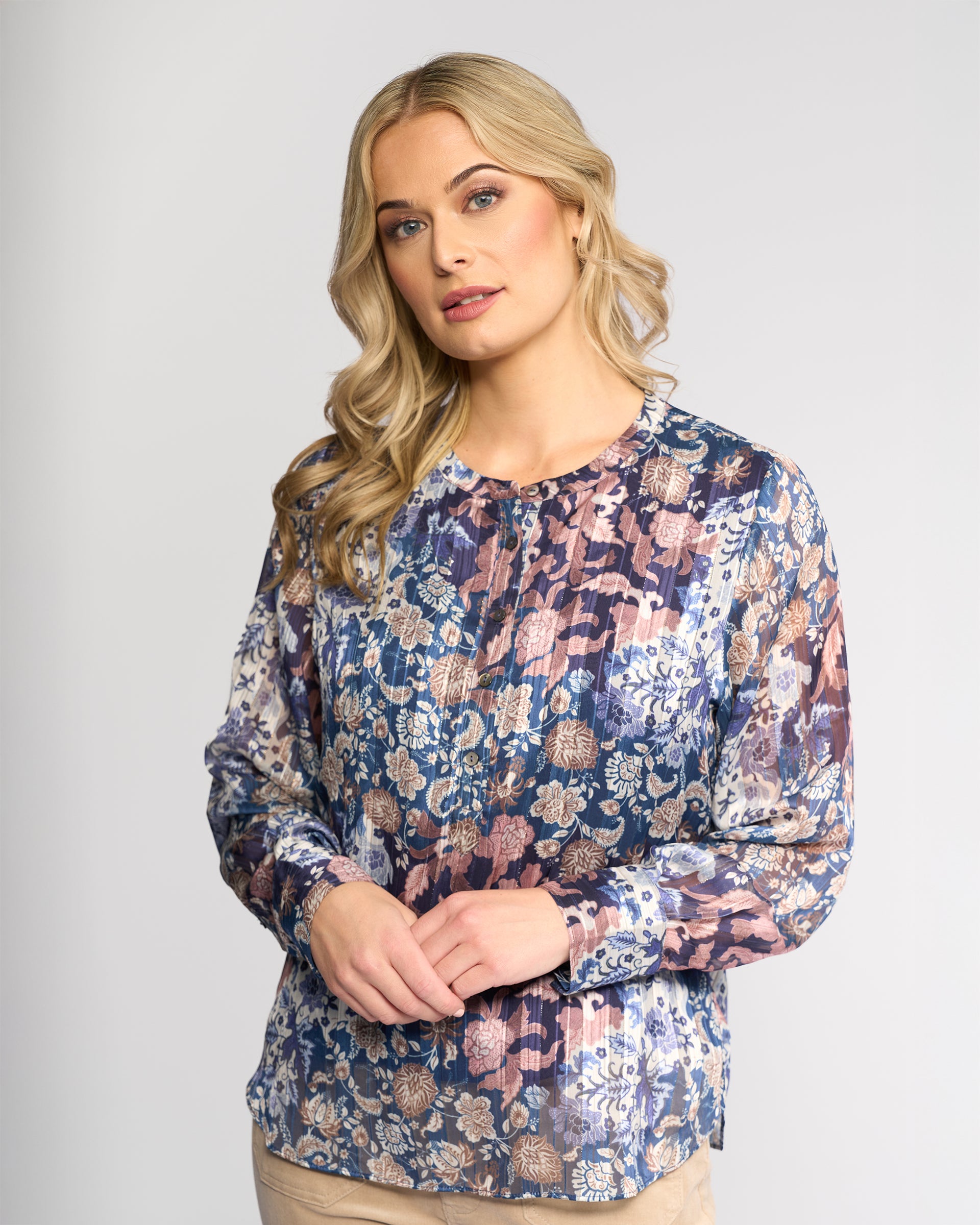 A woman with long blonde hair models the Vassalli Rhythm Top - Rhea, a long-sleeve chiffon shirt in blue, purple, and beige floral print. She stands against a light gray background, gazing slightly to the side with a neutral expression.