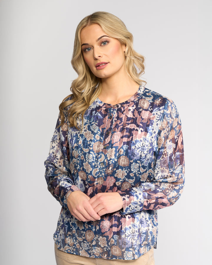 A woman with long blonde hair models the Vassalli Rhythm Top - Rhea, a long-sleeve chiffon shirt in blue, purple, and beige floral print. She stands against a light gray background, gazing slightly to the side with a neutral expression.