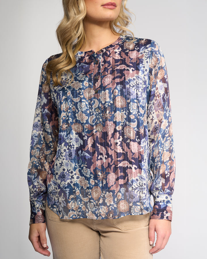 A woman wears the Vassalli Rhythm Top - Rhea, a long-sleeve chiffon shirt with a blue, purple, and beige floral print, paired with light tan pants. Her blonde hair falls in loose waves against a plain light background.