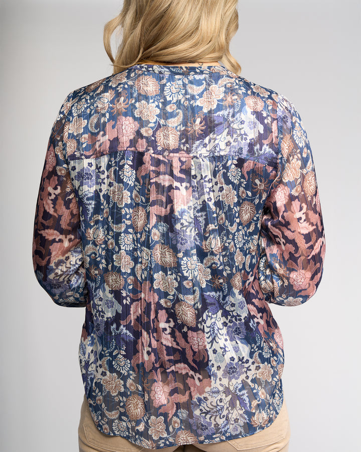 A person with blonde hair is pictured from the back wearing the Vassalli Rhythm Top - Rhea, a long-sleeve chiffon shirt featuring blue, pink, and beige floral prints, set against a plain light gray background.