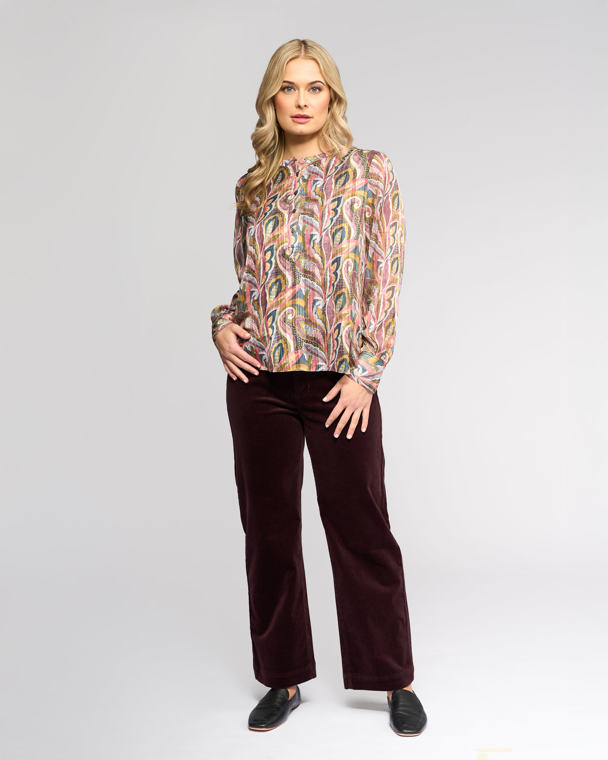 A woman with long blonde hair wears the Vassalli Rhythm Top - Sable and dark burgundy pants, standing against a plain light gray background while looking at the camera with a neutral expression.