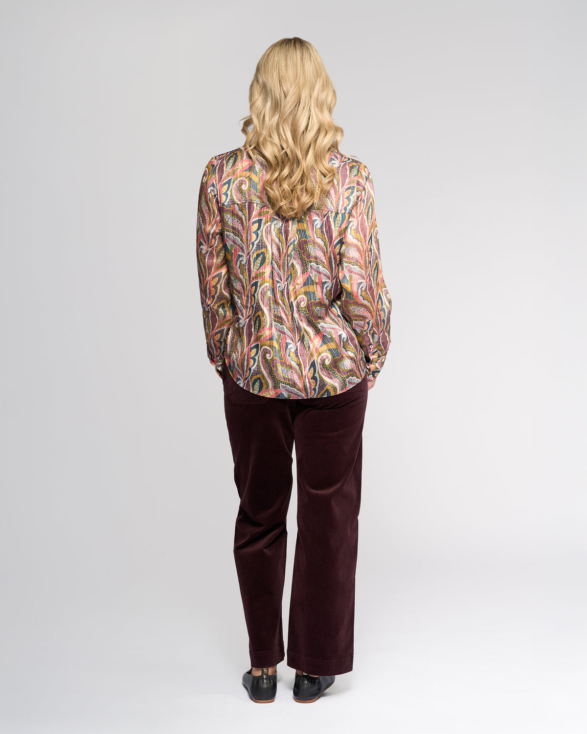 A person with long blonde hair, wearing the Vassalli Rhythm Top - Sable and dark pants, stands facing away from the camera against a plain light background.