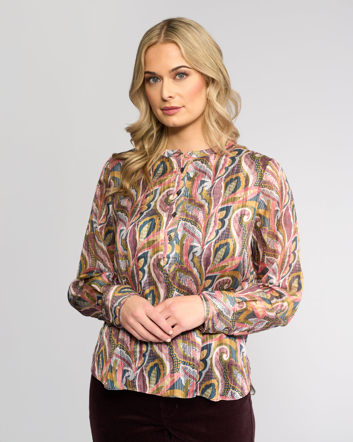 A woman with long blonde hair models the Vassalli Rhythm Top - Sable, a colorful paisley print long-sleeve blouse, paired with dark pants. She stands against a light grey background, looking slightly to the side with hands clasped.