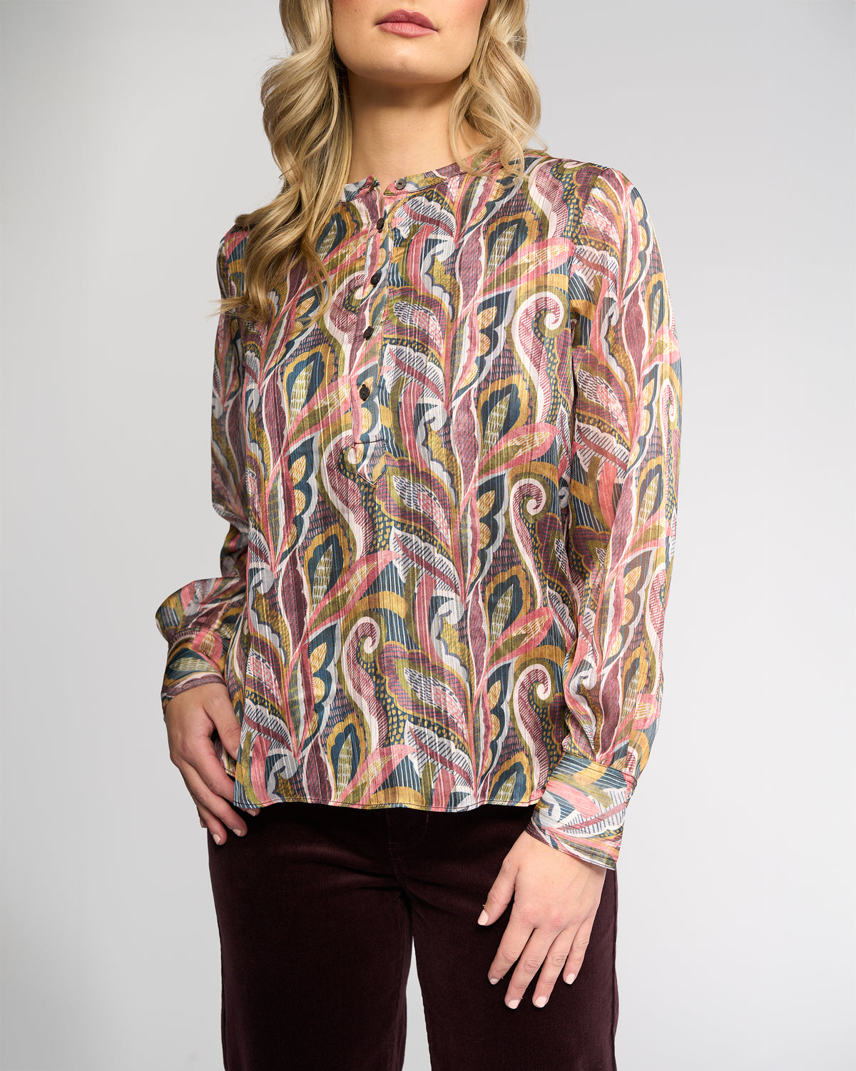 A woman stands against a plain light background wearing the Vassalli Rhythm Top - Sable and maroon pants. Her blonde hair is styled in loose waves, and her right hand rests on her hip.
