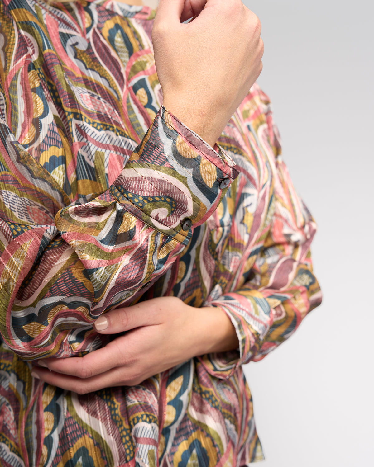 A person wears the Vassalli Rhythm Top - Sable, featuring a colorful paisley print and long sleeves. They cross one arm over their torso against a neutral backdrop; only arms and torso are visible.