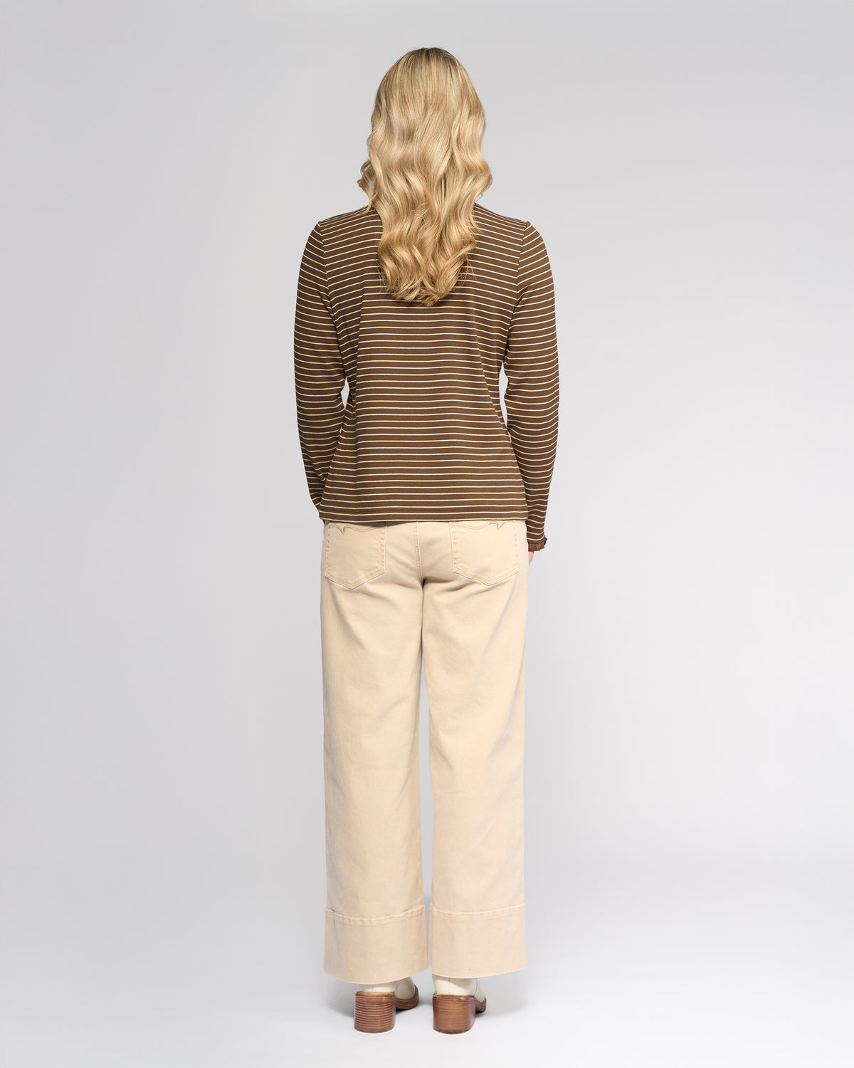 A woman with long, wavy blonde hair faces away from the camera, wearing the Vassalli Field Top in Chocolate/Oat with beige wide-leg pants and brown shoes against a plain light background.