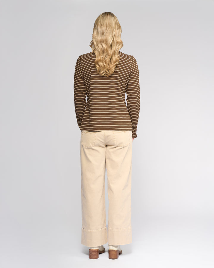 A woman with long, wavy blonde hair faces away from the camera, wearing the Vassalli Field Top in Chocolate/Oat with beige wide-leg pants and brown shoes against a plain light background.