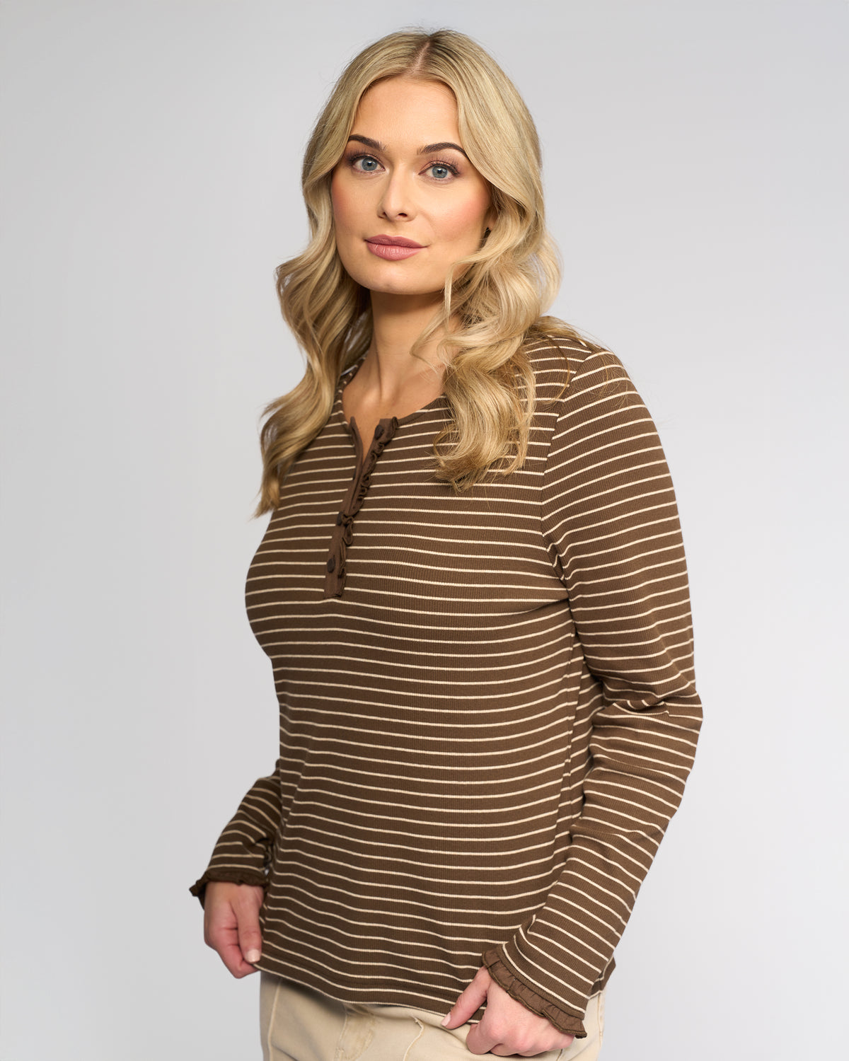 A woman with long blonde hair and light eyes models the Vassalli Field Top in Chocolate/Oat, featuring brown and white stripes, paired with beige pants against a plain light gray background. She gazes slightly aside with a neutral expression.