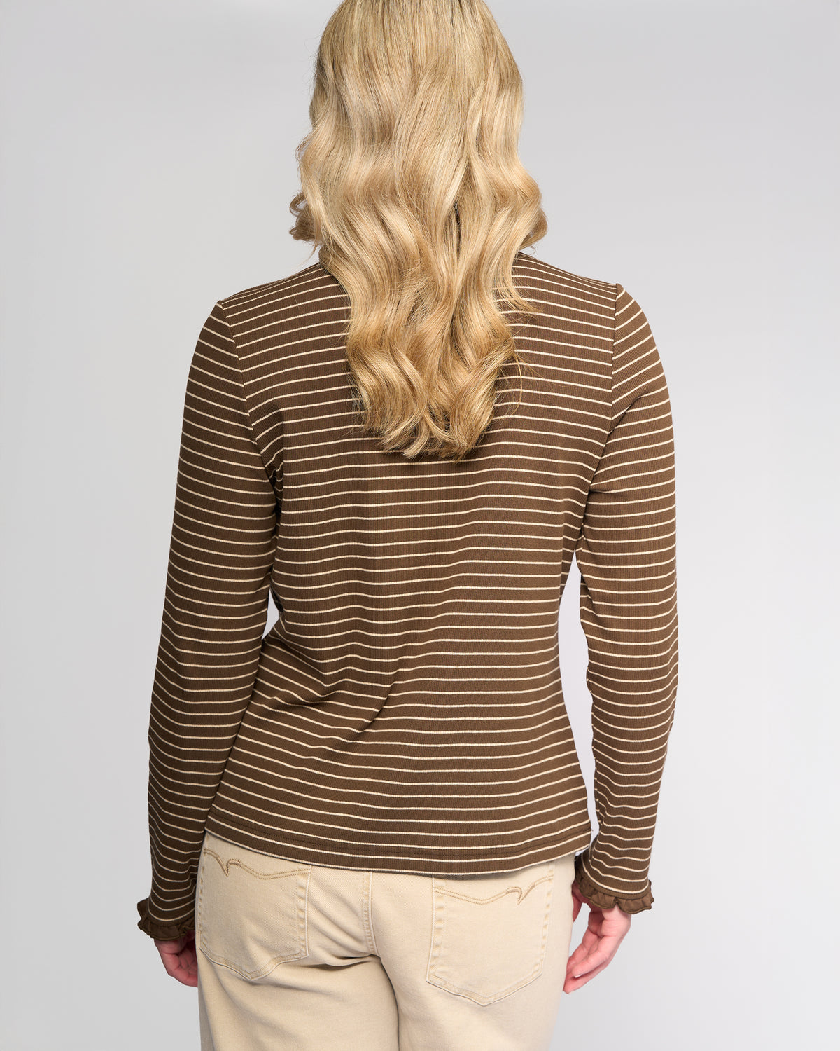 A person with long, wavy blonde hair is shown from the back wearing the Vassalli Field Top in Chocolate/Oat and light beige pants, against a plain background.