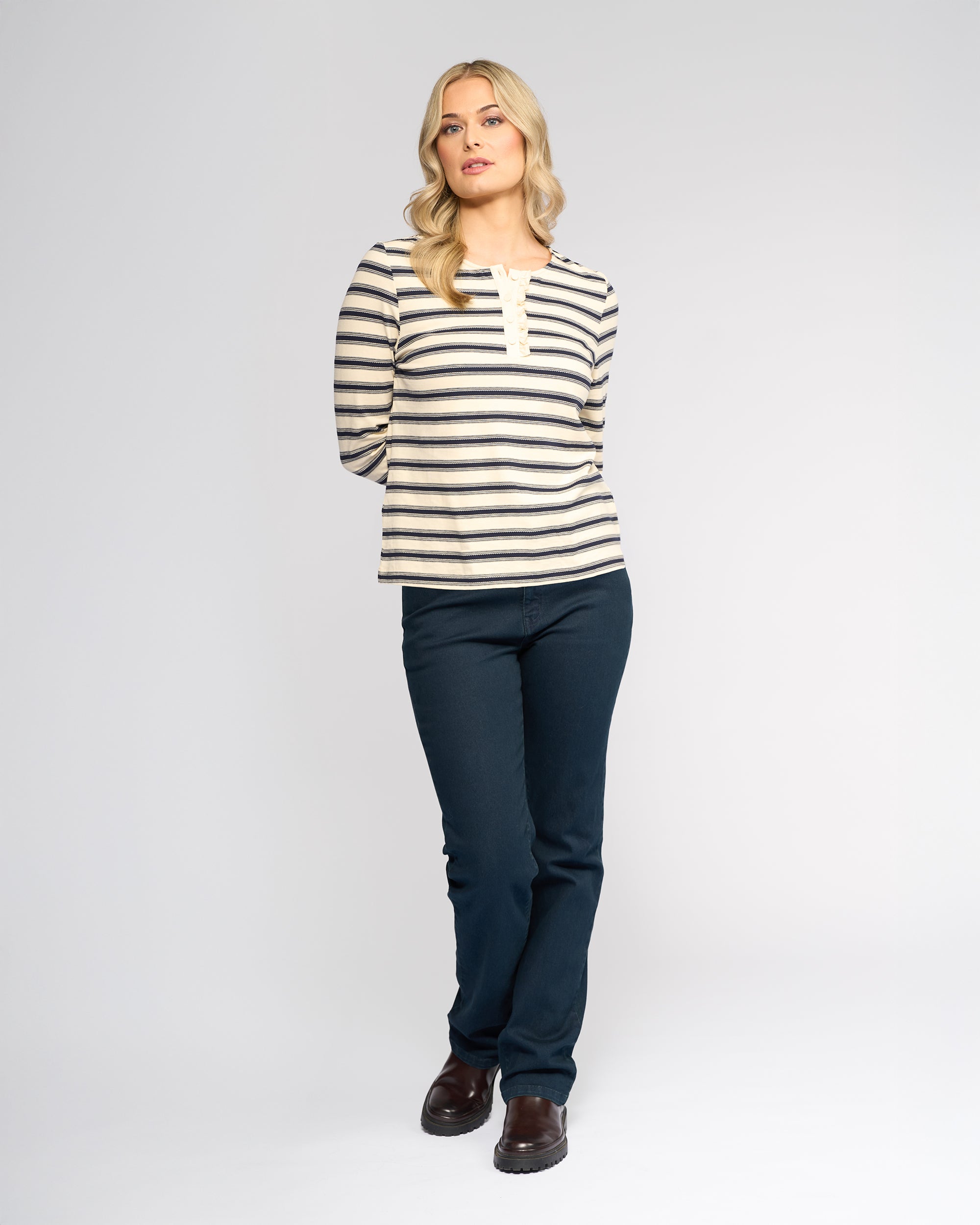 A woman with loose blond waves stands against a white background, wearing the Vassalli Field Top in Oat/Ink stripes, paired with dark blue pants and brown shoes. Her hands are behind her back.