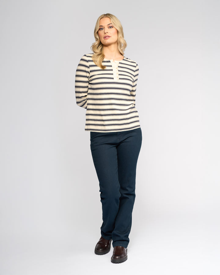 A woman with loose blond waves stands against a white background, wearing the Vassalli Field Top in Oat/Ink stripes, paired with dark blue pants and brown shoes. Her hands are behind her back.