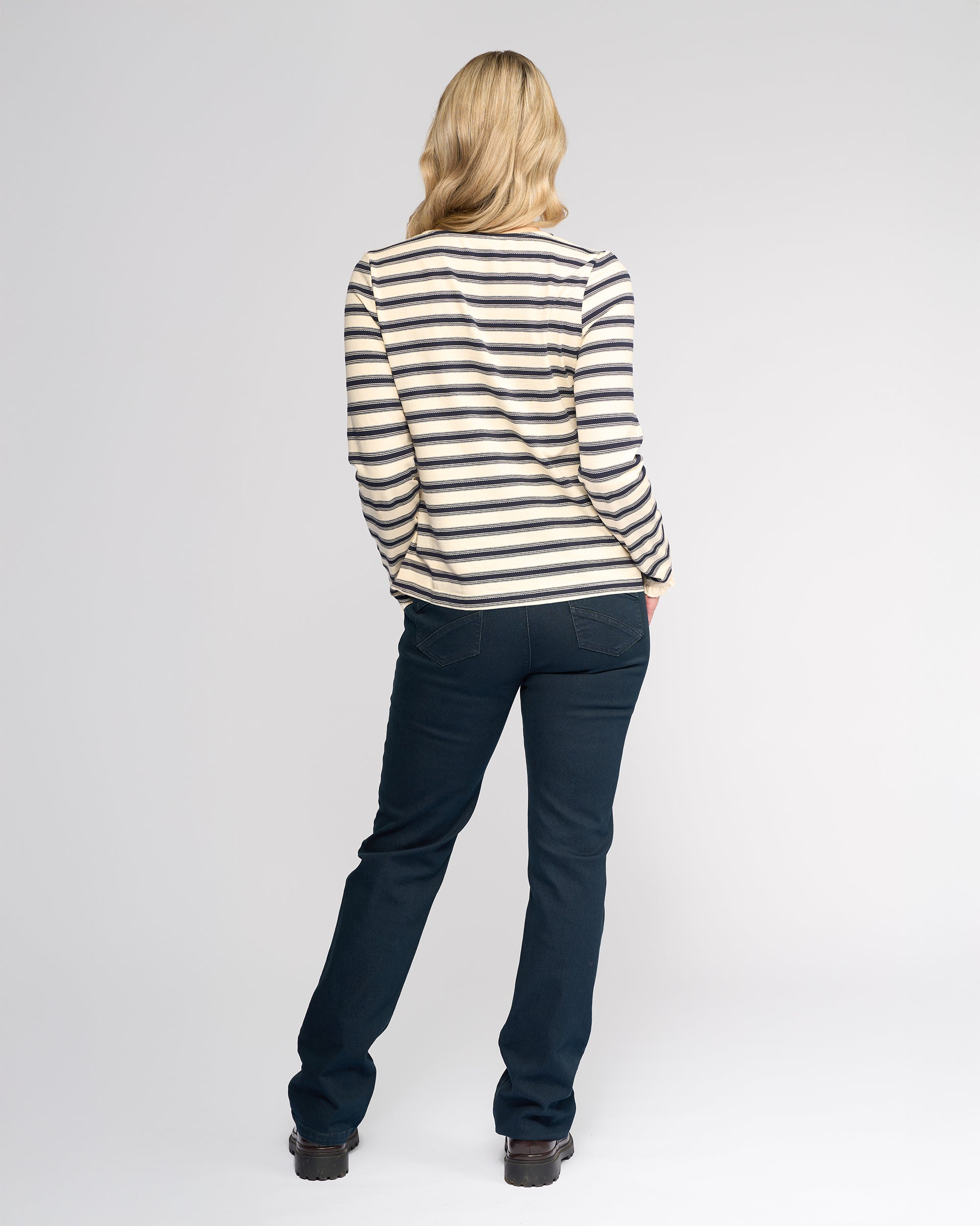 A person with shoulder-length blonde hair stands facing away, wearing the Vassalli Field Top in Oat/Ink, dark blue jeans, and black shoes against a plain white background.