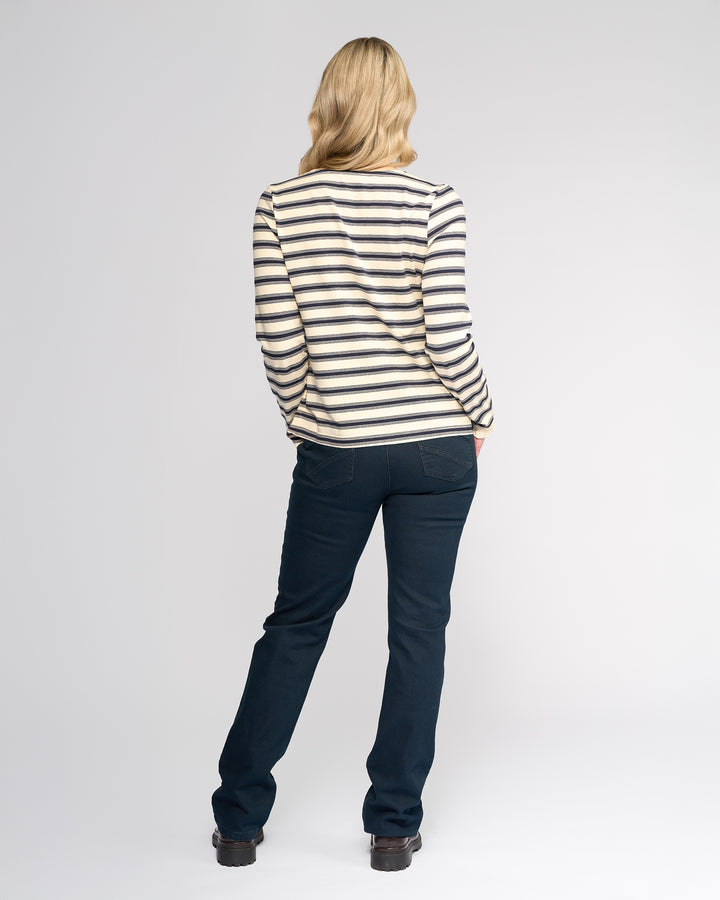 A person with shoulder-length blonde hair stands facing away, wearing the Vassalli Field Top in Oat/Ink, dark blue jeans, and black shoes against a plain white background.