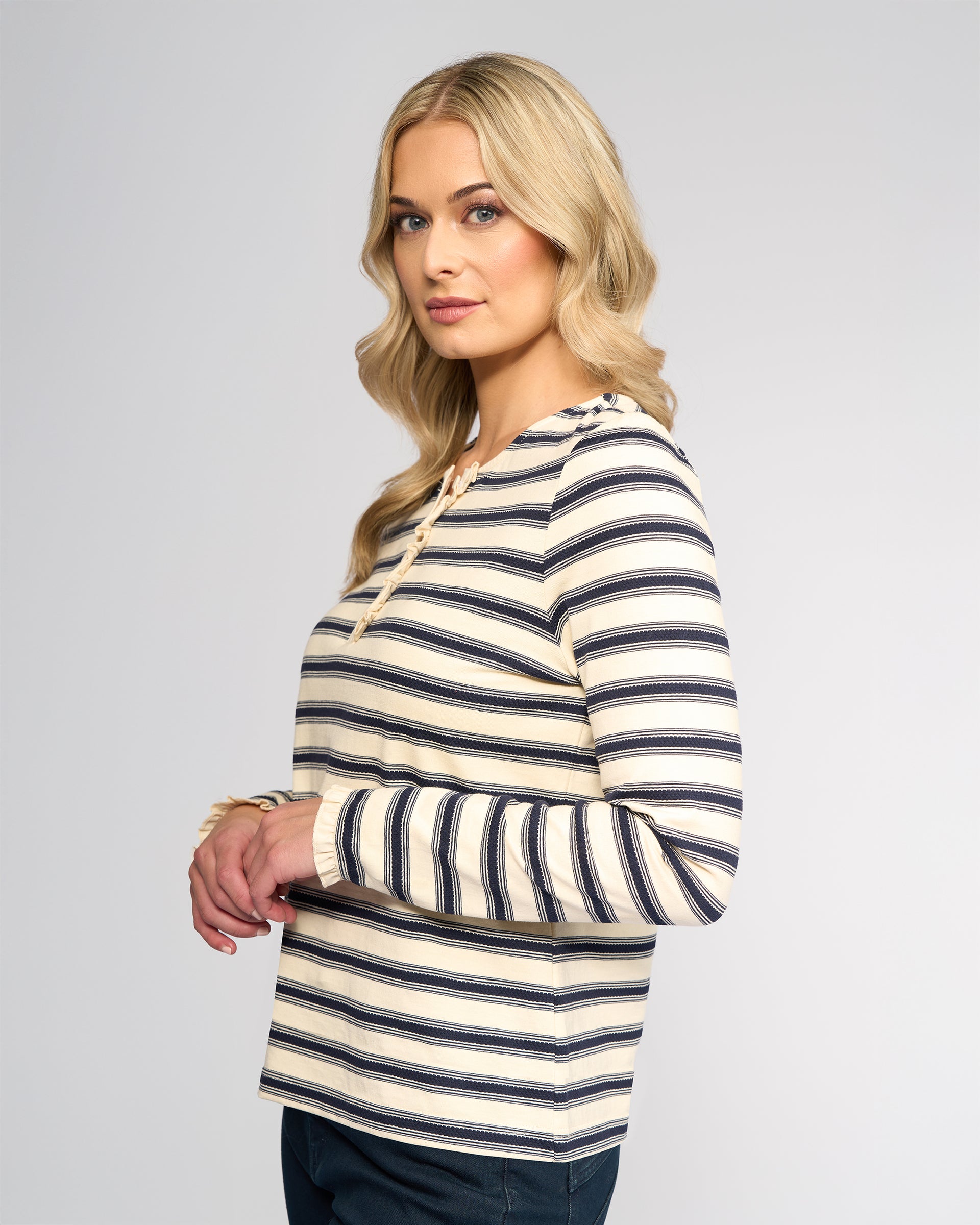 A woman with long blonde hair stands sideways, wearing Vassalli's Field Top in Oat/Ink, a cream and navy striped long-sleeve with subtle ruffle detail, looking neutrally at the camera against a plain background.