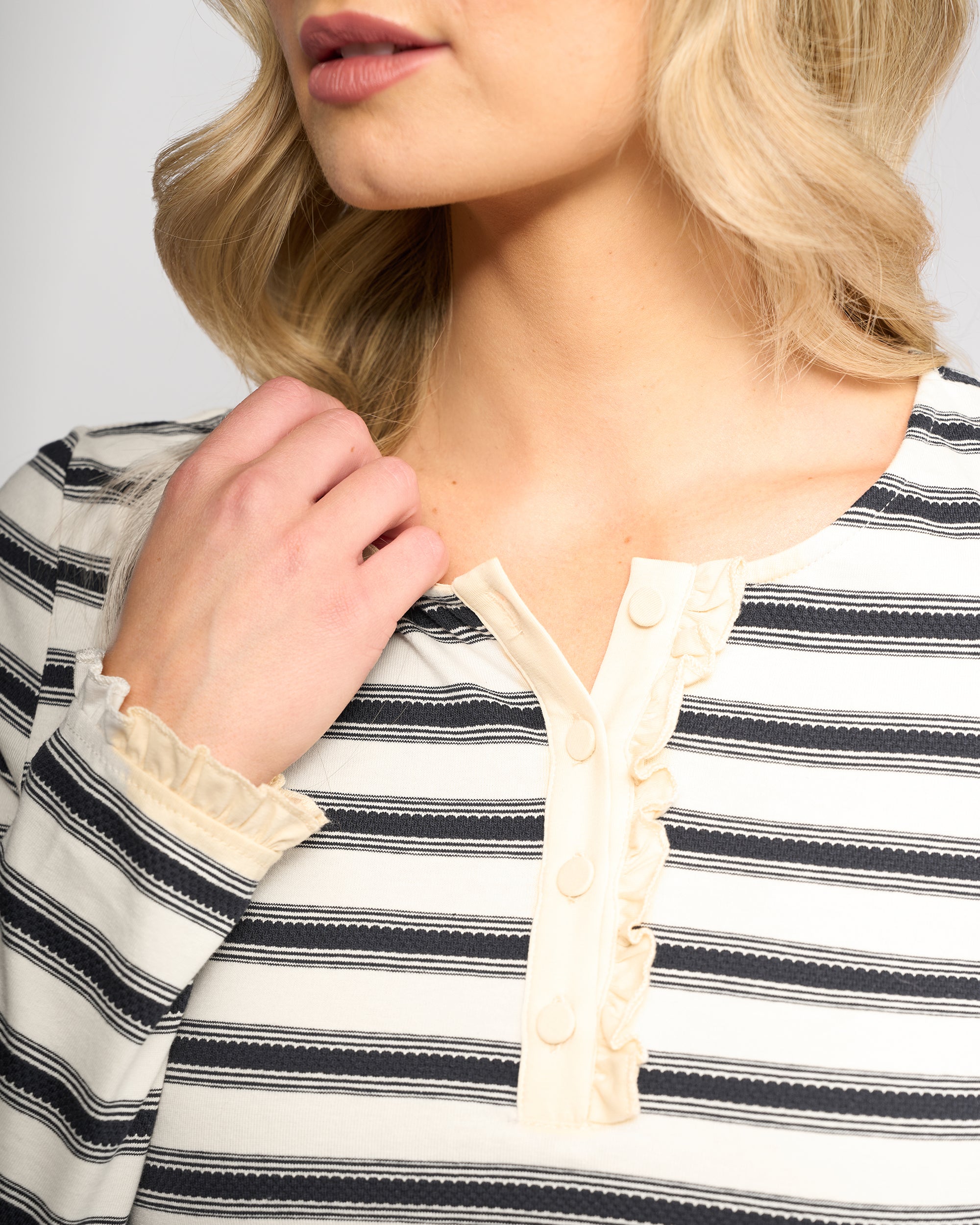 A woman with wavy blonde hair, her face partly visible and hand in her hair, wears the Vassalli Field Top - Oat/Ink: a black and white striped long-sleeve top with cream ruffle detailing and buttons on the collar.