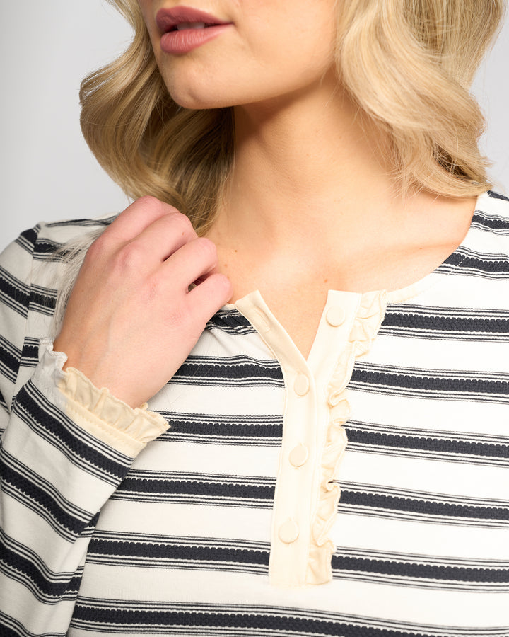 A woman with wavy blonde hair, her face partly visible and hand in her hair, wears the Vassalli Field Top - Oat/Ink: a black and white striped long-sleeve top with cream ruffle detailing and buttons on the collar.