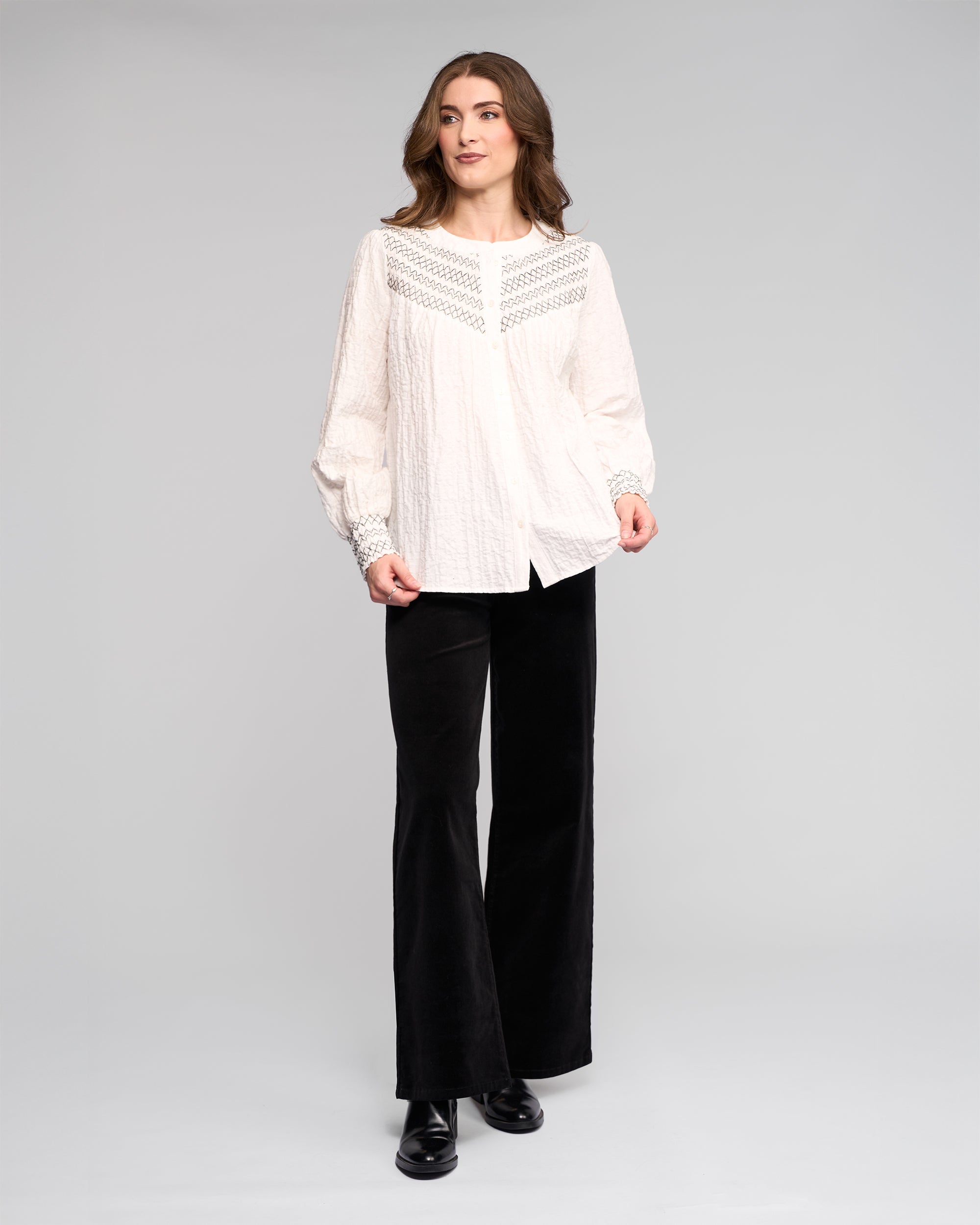A woman with wavy brown hair and a neutral expression wears the Vassalli Charm Shirt in off white, featuring lace and black stitched details, paired with black flared pants and matching shoes against a plain background.