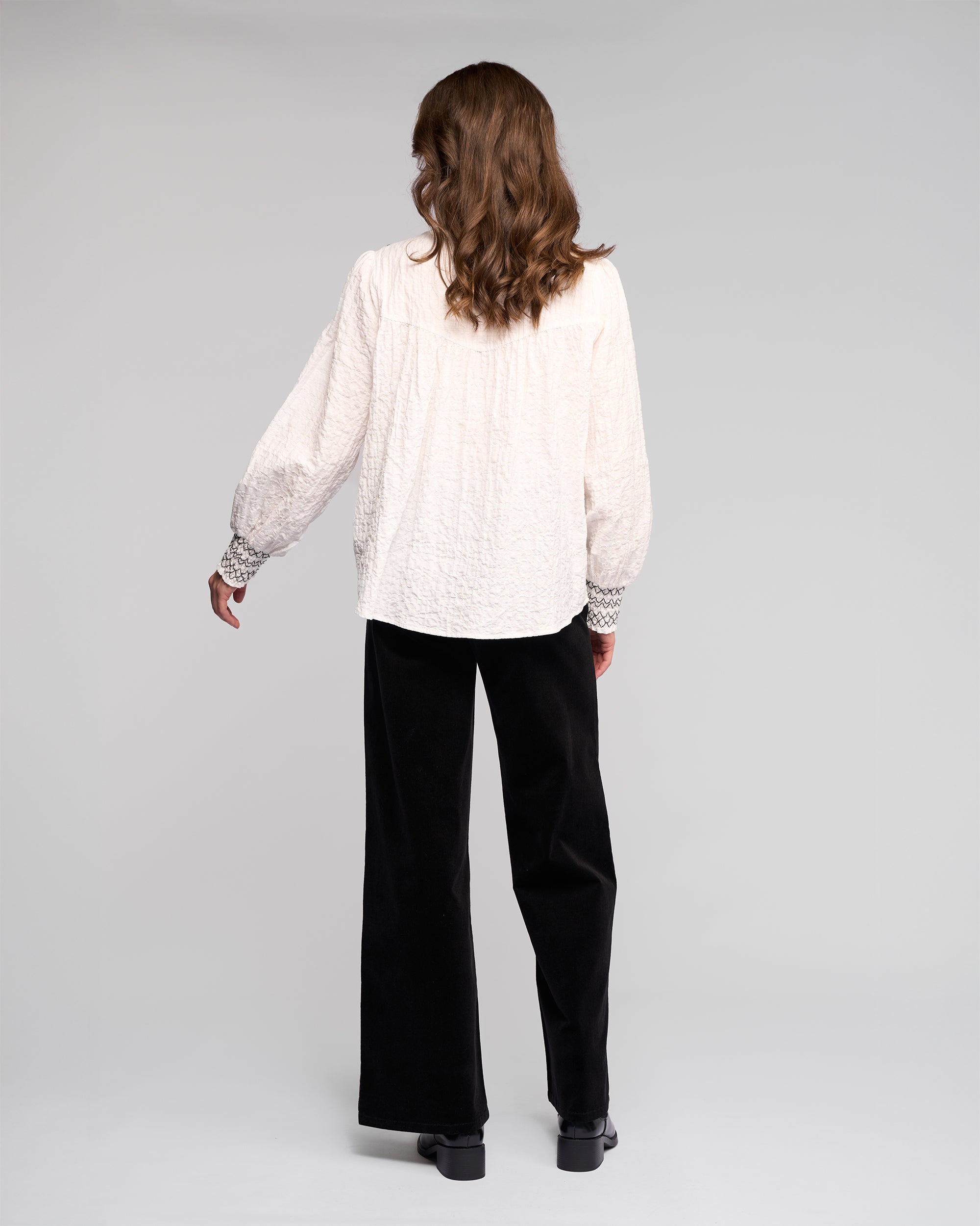 A person with wavy brown hair, seen from behind, wears the Vassalli Charm Shirt in off white with black stitched details and loose sleeves, paired with black wide-leg pants against a plain light background.
