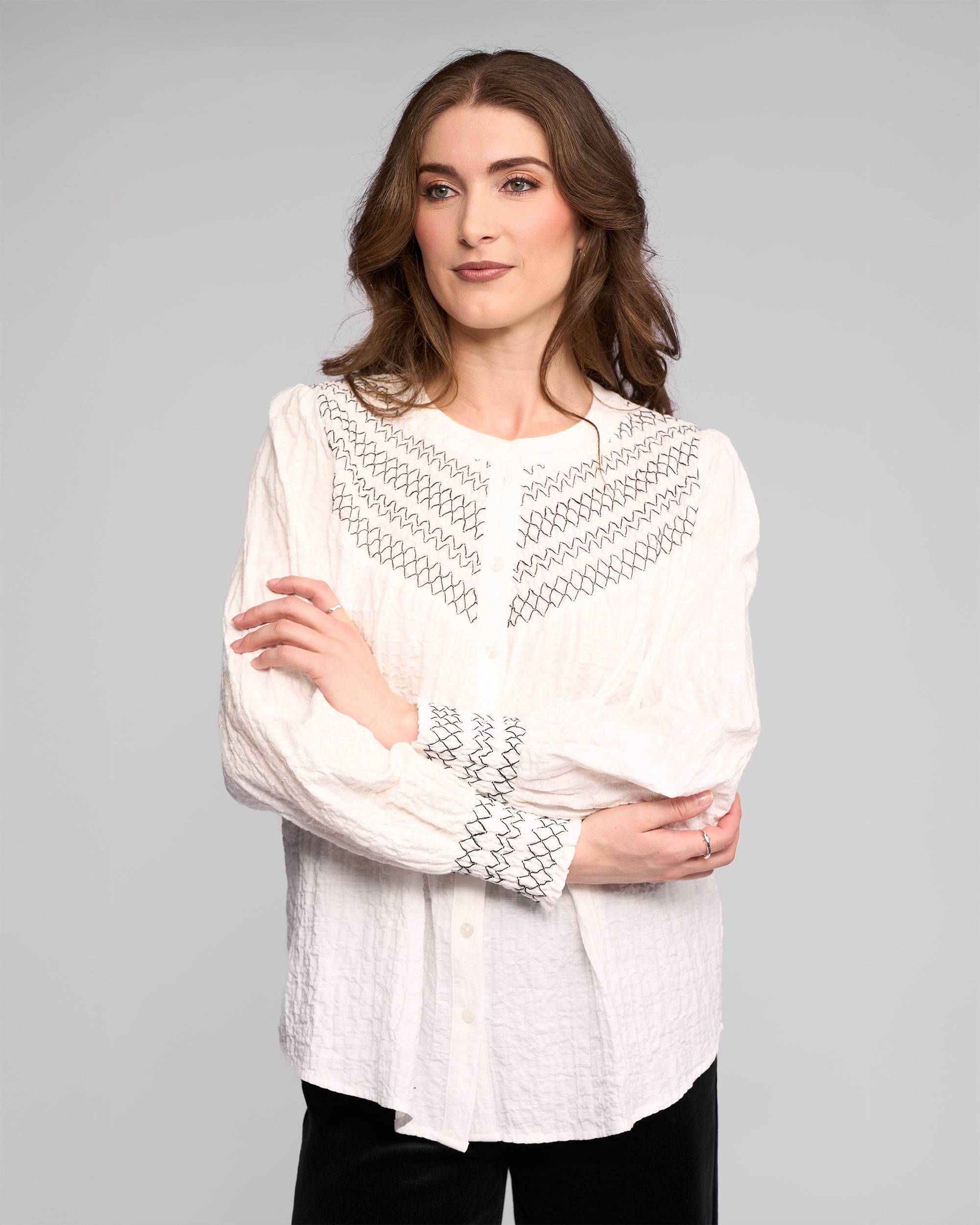 A woman with long brown hair models the Vassalli Charm Shirt in off white, featuring black stitched detailing. She stands against a light gray background, looking to the side with a relaxed expression and arms loosely crossed.