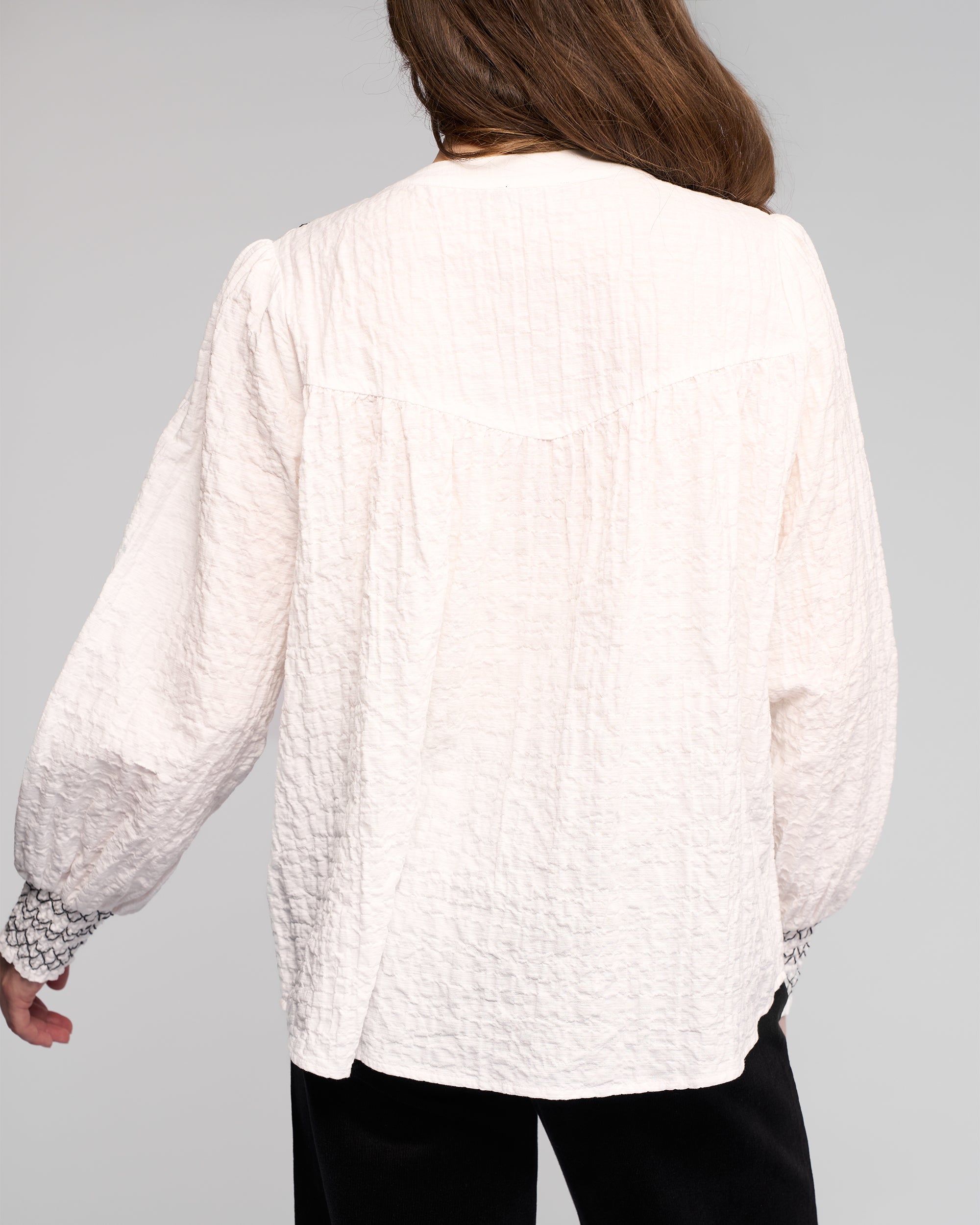 A person with long brown hair is seen from the back, wearing the Vassalli Charm Shirt in off white featuring black stitched cuffs, paired with black pants, against a plain light gray background.