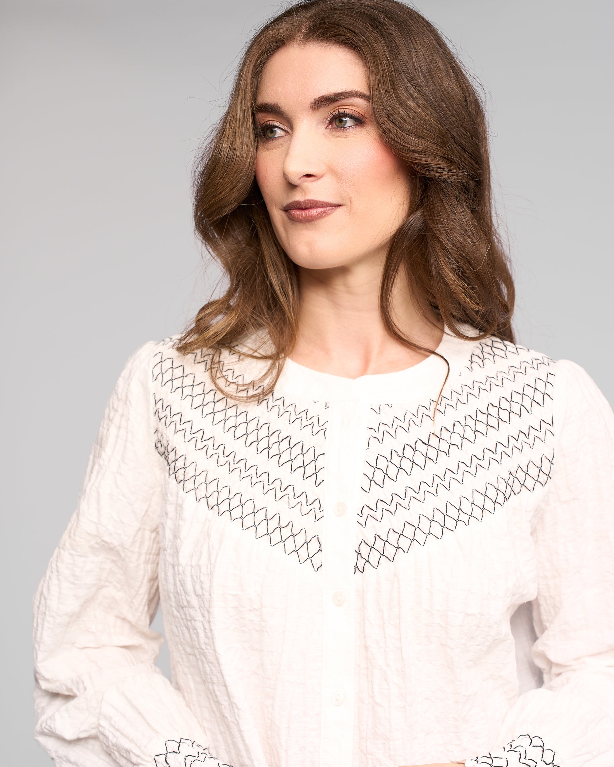 A woman with long brown hair, wearing the Vassalli Charm Shirt in off white—featuring black zigzag and diamond patterns plus stitched chest and cuff details—looks to the side against a light gray background.