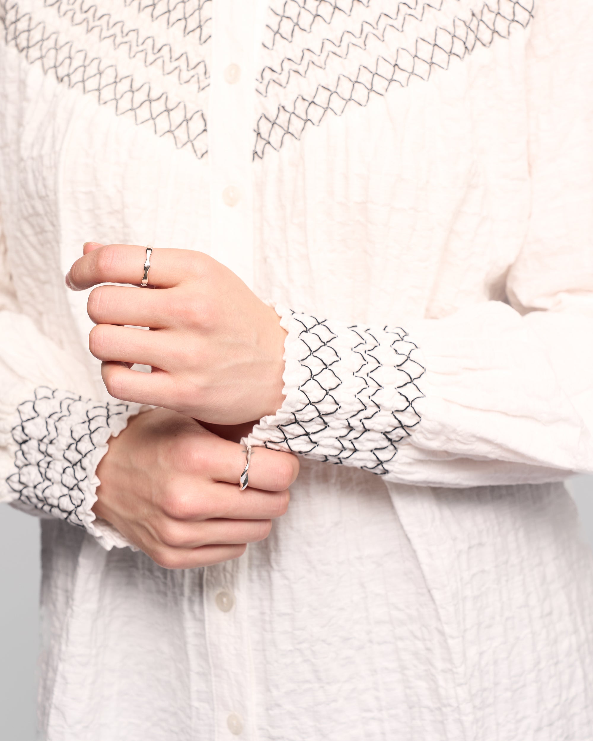 Wearing the Vassalli Charm Shirt in off white, which features black zigzag embroidery on the cuffs and upper chest, a person lifts one sleeve to display rings on their fingers.