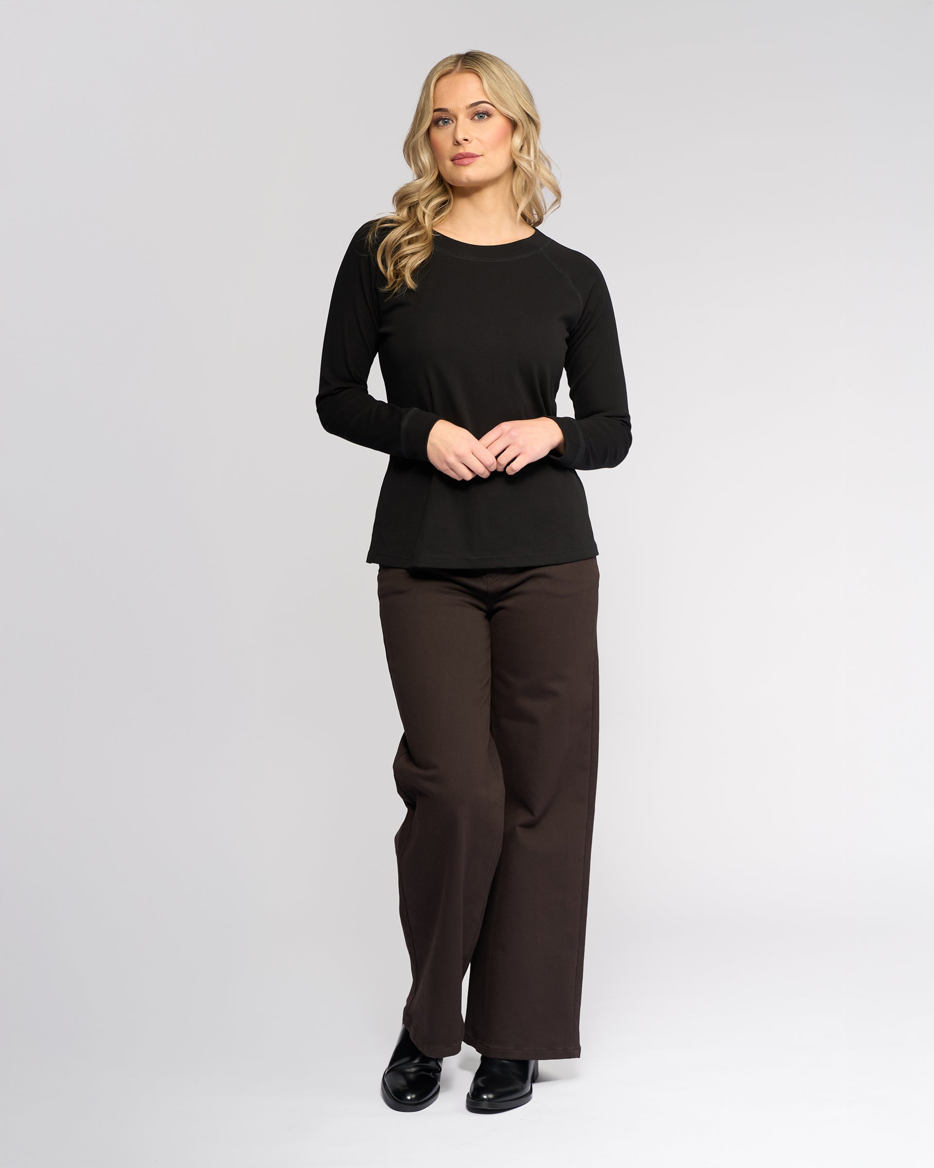 A woman with long blonde hair wears the Vassalli Key Top in black, paired with brown wide-leg pants and black shoes, standing against a plain light gray background.