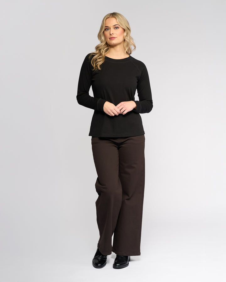 A woman with long blonde hair wears the Vassalli Key Top in black, paired with brown wide-leg pants and black shoes, standing against a plain light gray background.
