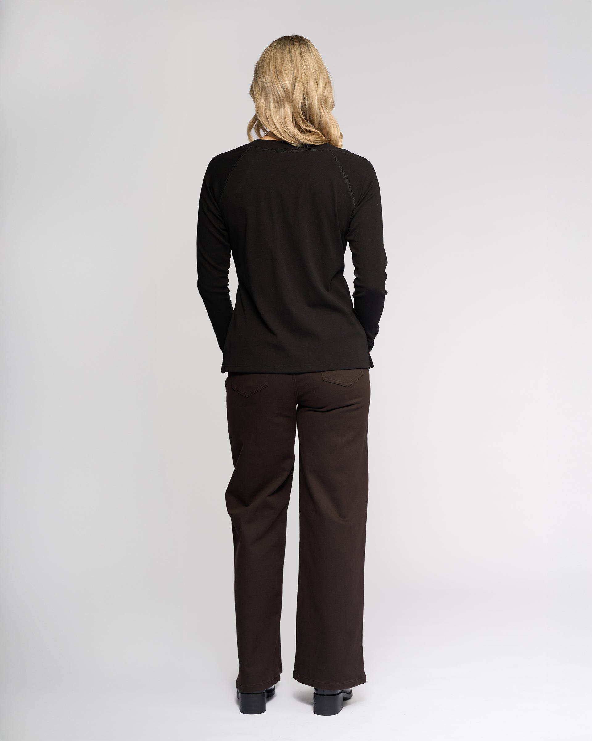 A person with long blonde hair, seen from behind, wears the Vassalli Key Top in black with dark brown pants and black shoes against a plain light background.