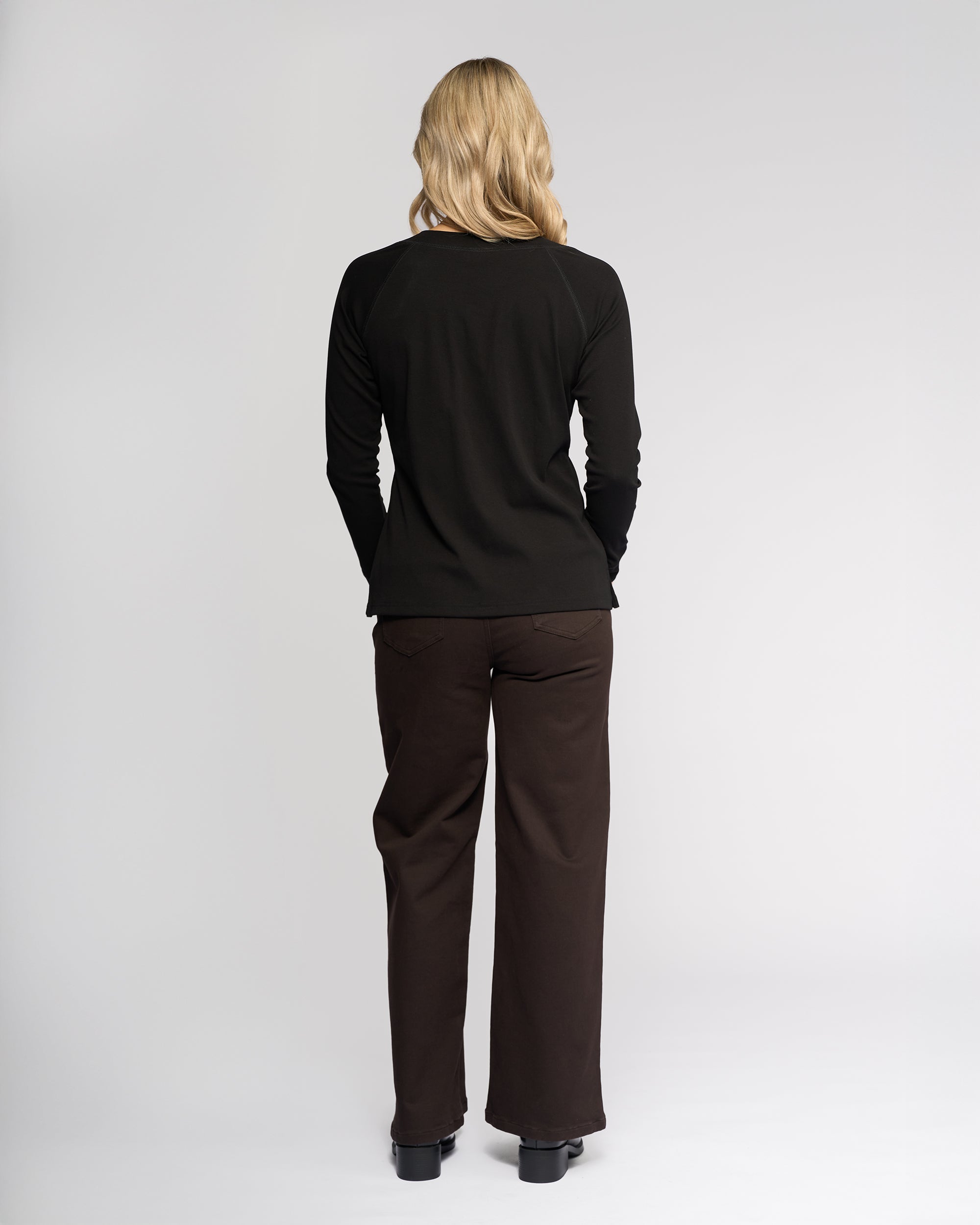 A person with long blonde hair, seen from behind, wears the Vassalli Key Top in black with dark brown pants and black shoes against a plain light background.