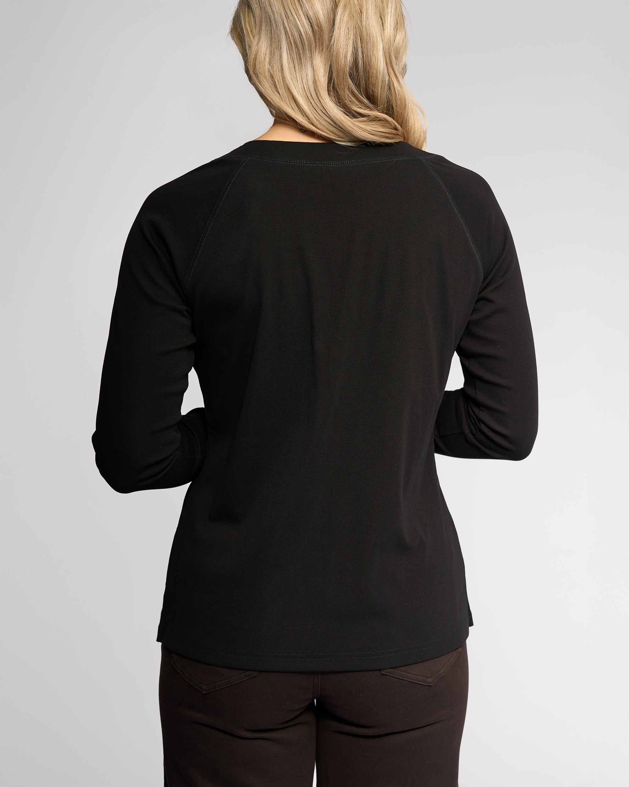 A person with shoulder-length blonde hair is shown from the back, wearing the Vassalli Key Top - Black and dark pants against a plain light background.