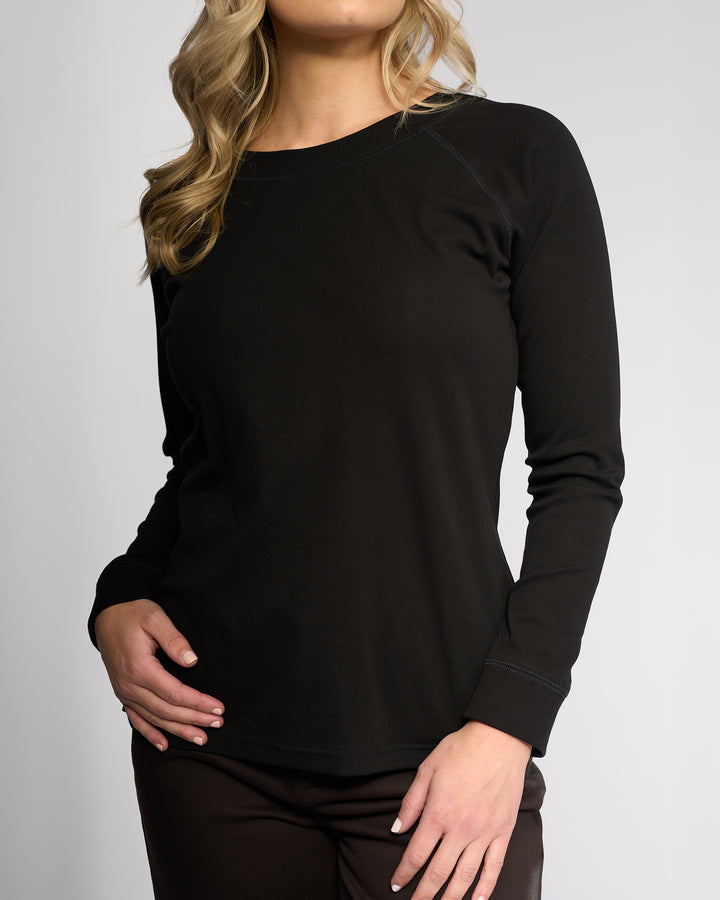 A woman with wavy blonde hair models the Vassalli Key Top in black, paired with dark pants. She poses with one hand on her hip against a plain light background, her face partially out of frame.