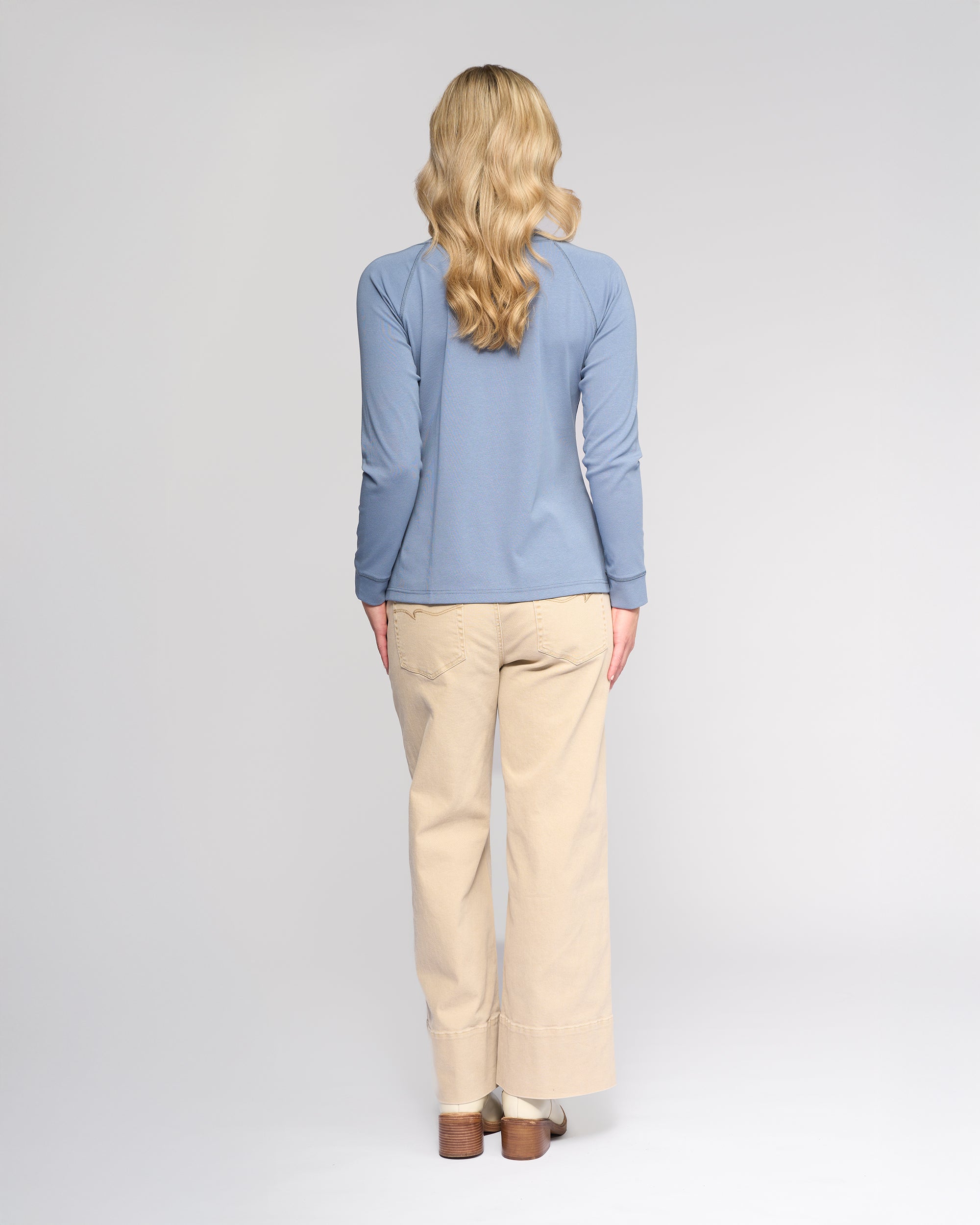 A person with long, wavy blonde hair stands facing away, wearing the Vassalli Key Top in Denim Blue, beige wide-leg pants, and brown shoes against a plain light background.