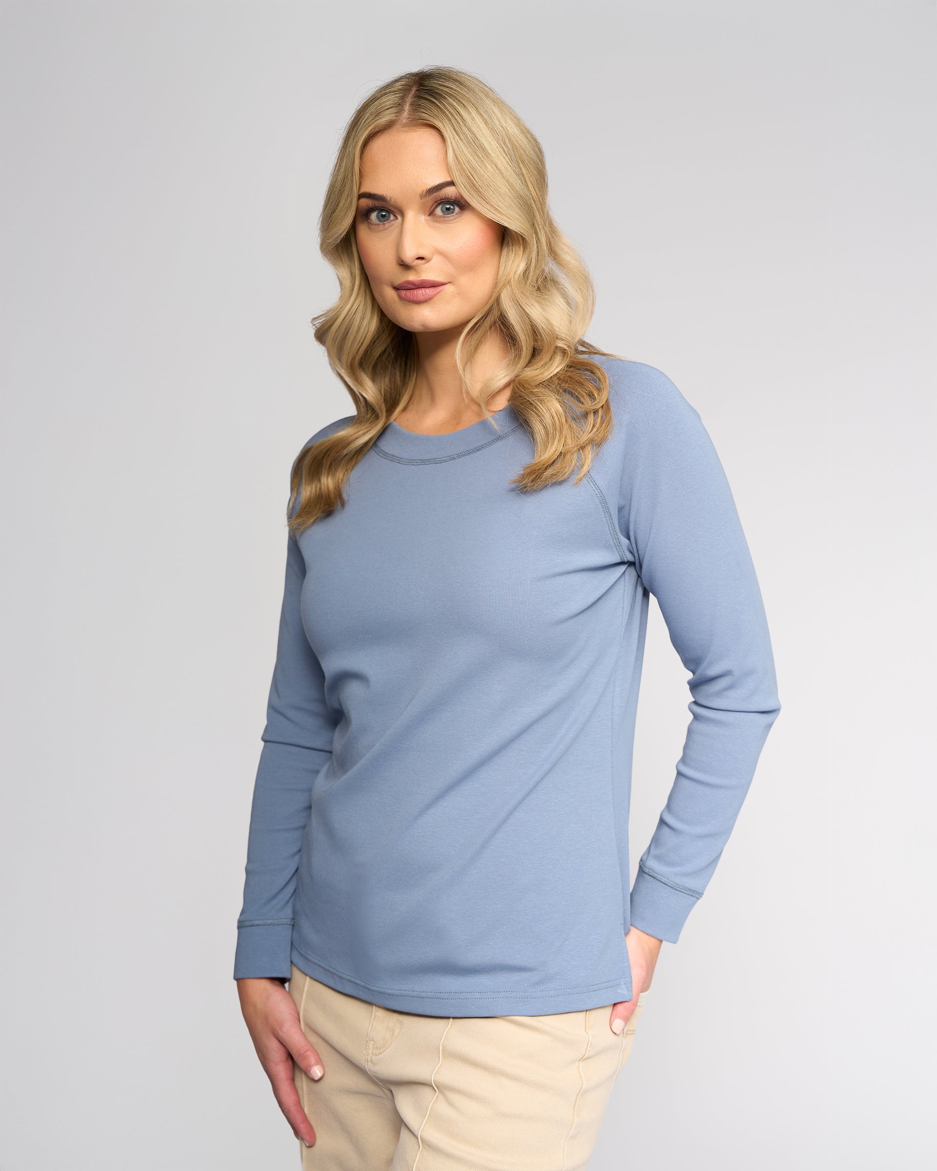 A woman with long blonde hair wears the Vassalli Key Top in denim blue, paired with beige pants, standing against a plain light gray background and looking slightly to the side with a neutral expression.