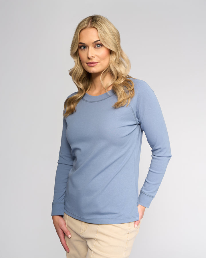 A woman with long blonde hair wears the Vassalli Key Top in denim blue, paired with beige pants, standing against a plain light gray background and looking slightly to the side with a neutral expression.