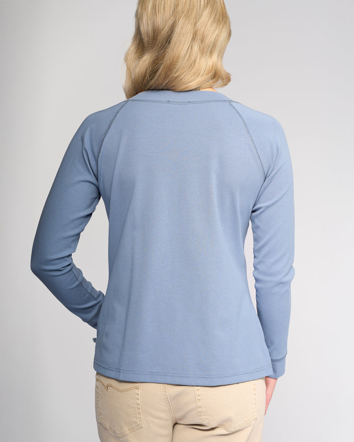 A person with blonde hair is facing away, wearing the Vassalli Key Top in denim blue and light beige pants, against a plain light background.