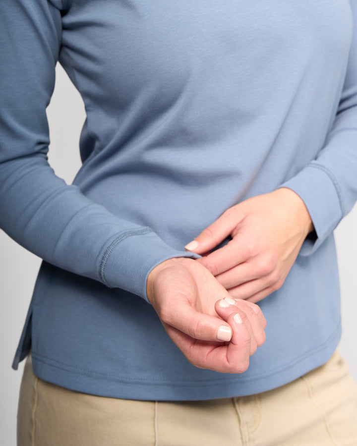 A person models the Vassalli Key Top in denim blue, standing against a plain background with their face hidden, and gently holding one wrist with the other hand.