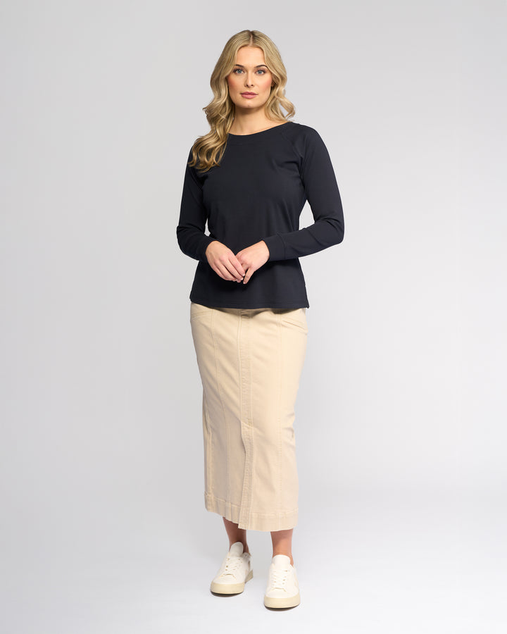 A woman with long blonde hair stands against a plain light background, wearing the Vassalli Key Top in Ink, a beige midi skirt, and white sneakers. She looks forward, hands loosely clasped, for a casual yet polished look.