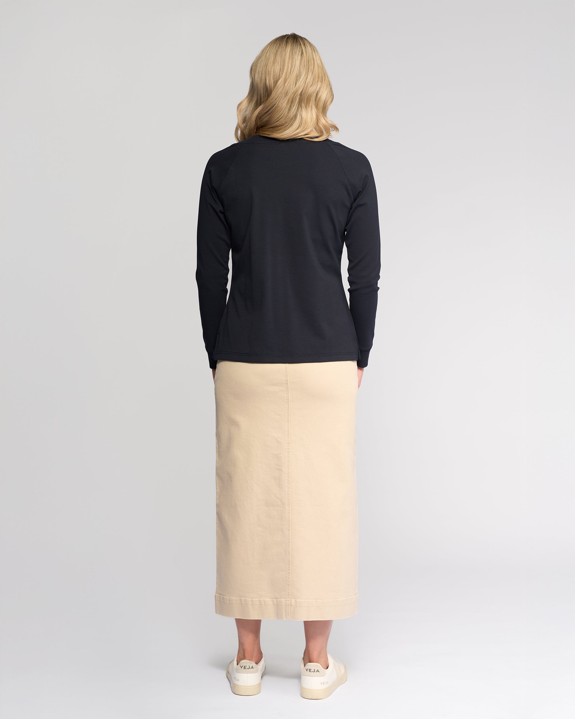 A woman faces away, wearing the Vassalli Key Top in Ink with a beige ankle-length skirt and white sneakers against a plain light background.