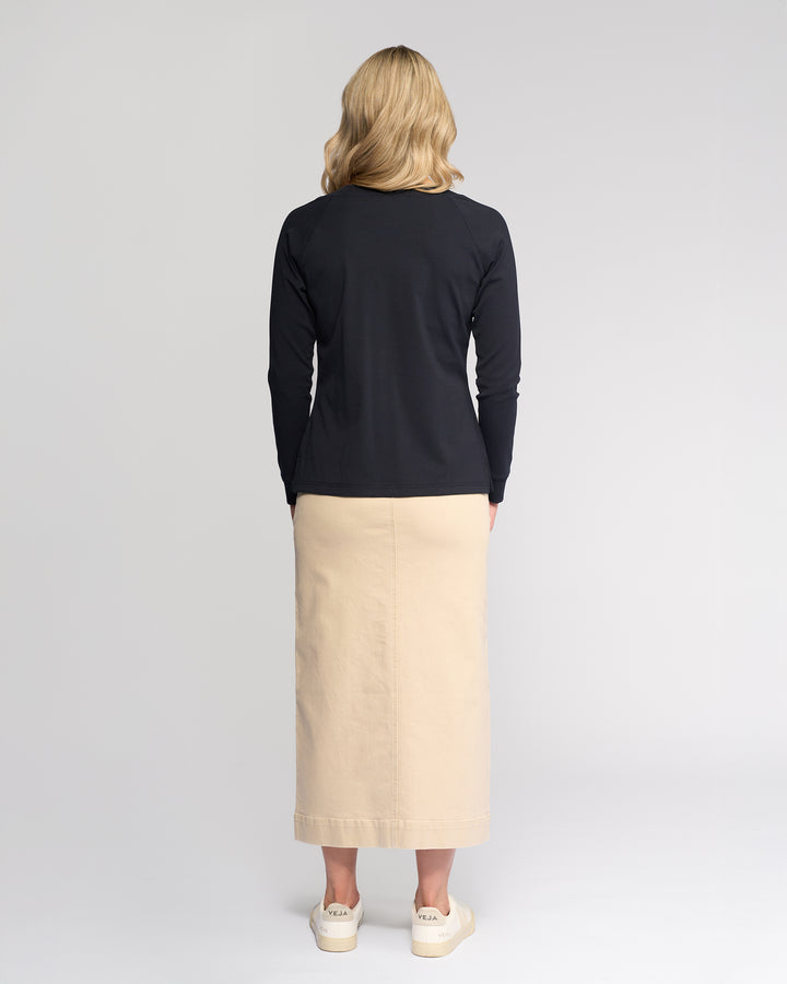 A woman faces away, wearing the Vassalli Key Top in Ink with a beige ankle-length skirt and white sneakers against a plain light background.
