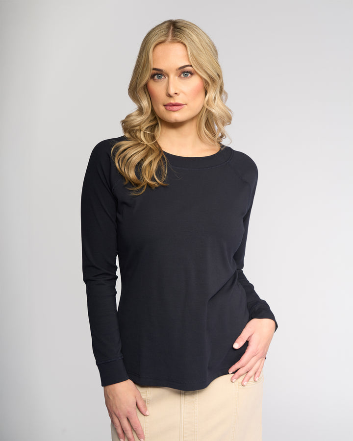 A woman with long blonde hair wears the Vassalli Key Top - Ink, a casual black long-sleeve stretch cotton top, paired with a beige skirt, standing against a light gray background and looking directly at the camera.