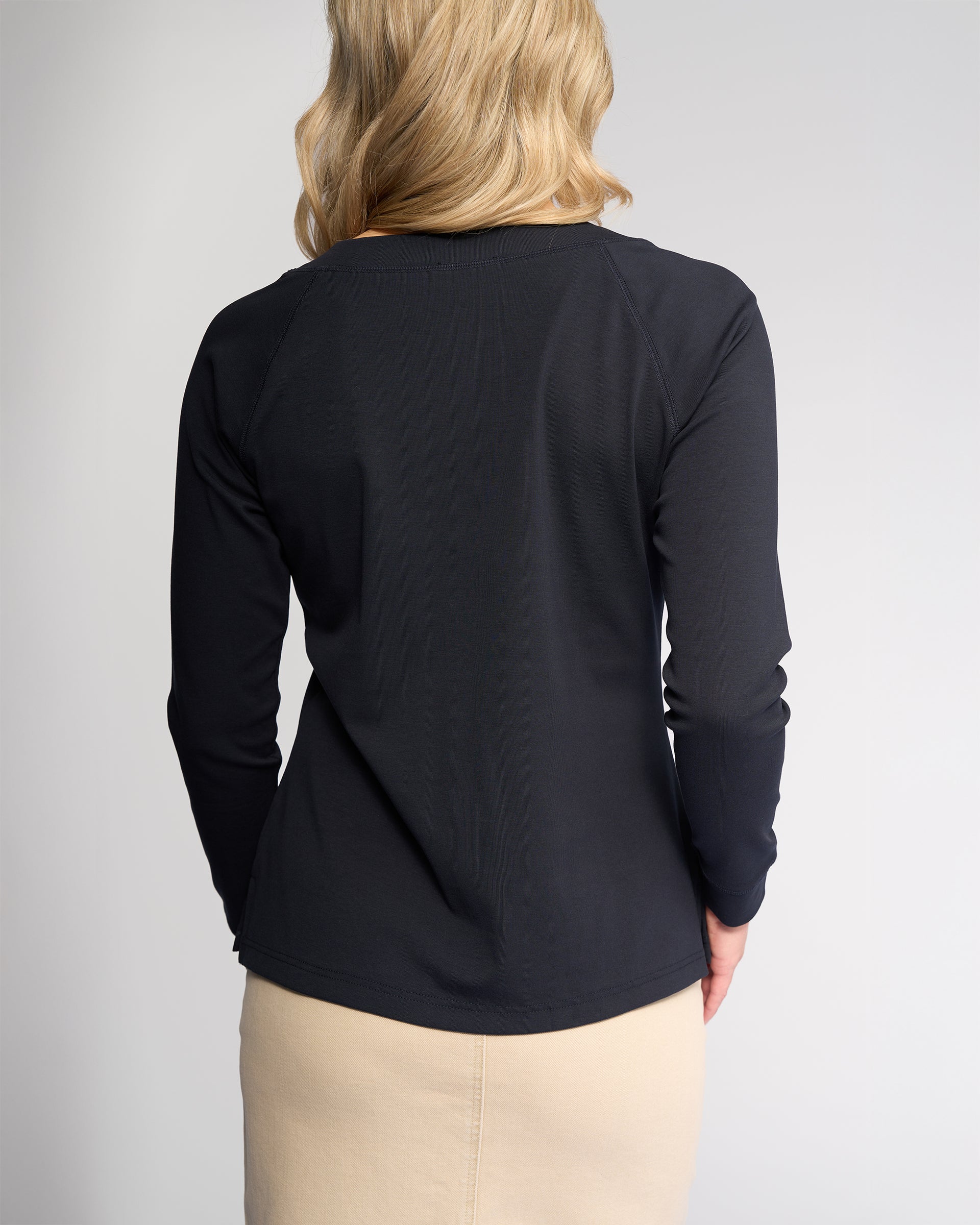 A woman with blonde, wavy hair is shown from behind wearing the Vassalli Key Top in Ink and a beige stretch cotton skirt, set against a plain light gray background.