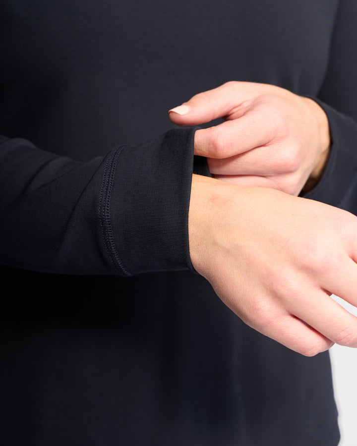 A close-up of hands adjusting the sleeve of the Vassalli Key Top - Ink, a black stretch cotton long-sleeve, with part of the torso visible against a plain background for a relaxed, casual look.