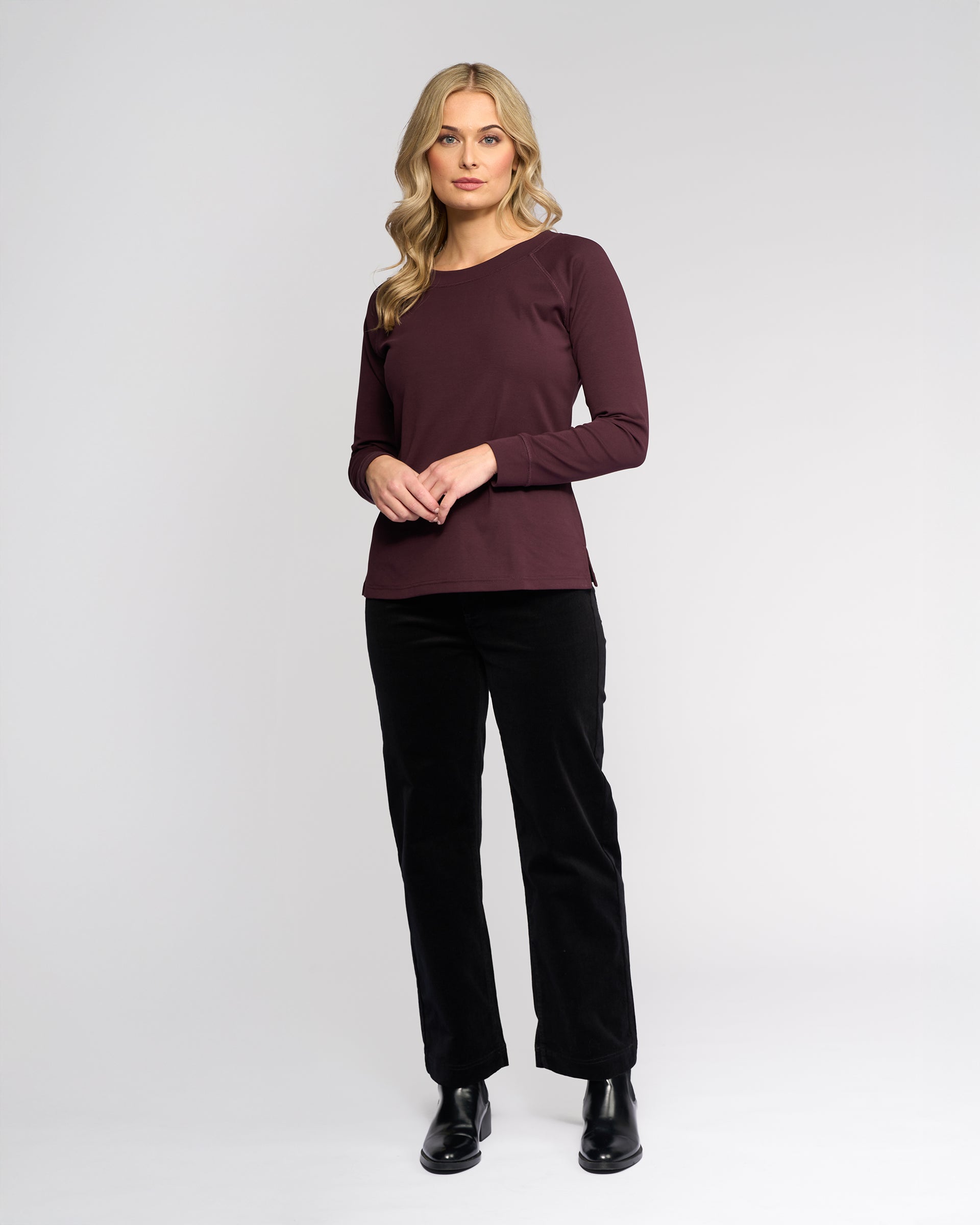 A woman with long blonde hair stands against a plain background wearing the Vassalli Key Top in Pinot, paired with black pants and boots. She has a neutral expression and her hands are lightly clasped.